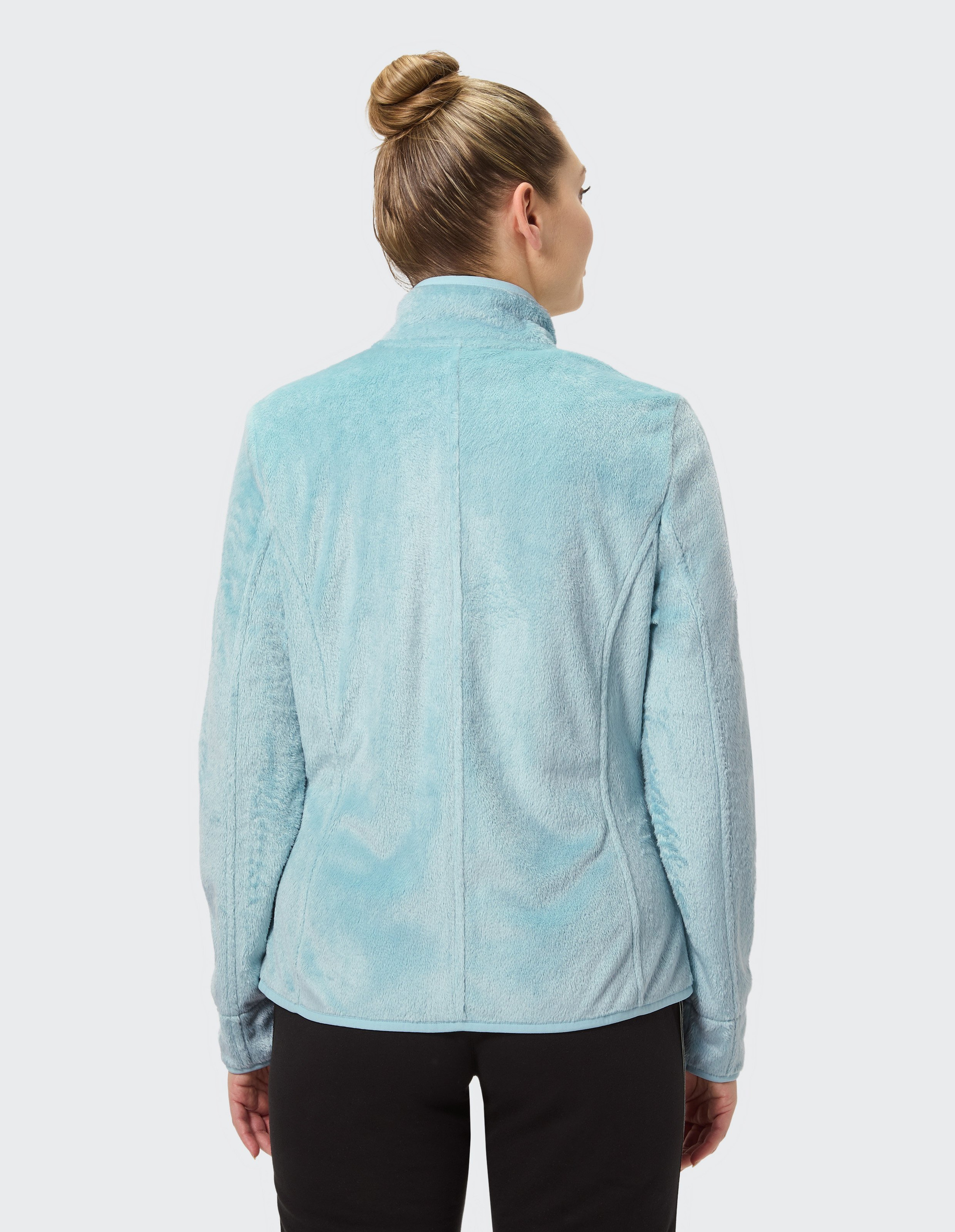 Frau mit Dutt von hinten in hellblauer Fleecejacke mit hohem Kragen, schwarzen Hosen, auf grauem Hintergrund betrachtet.