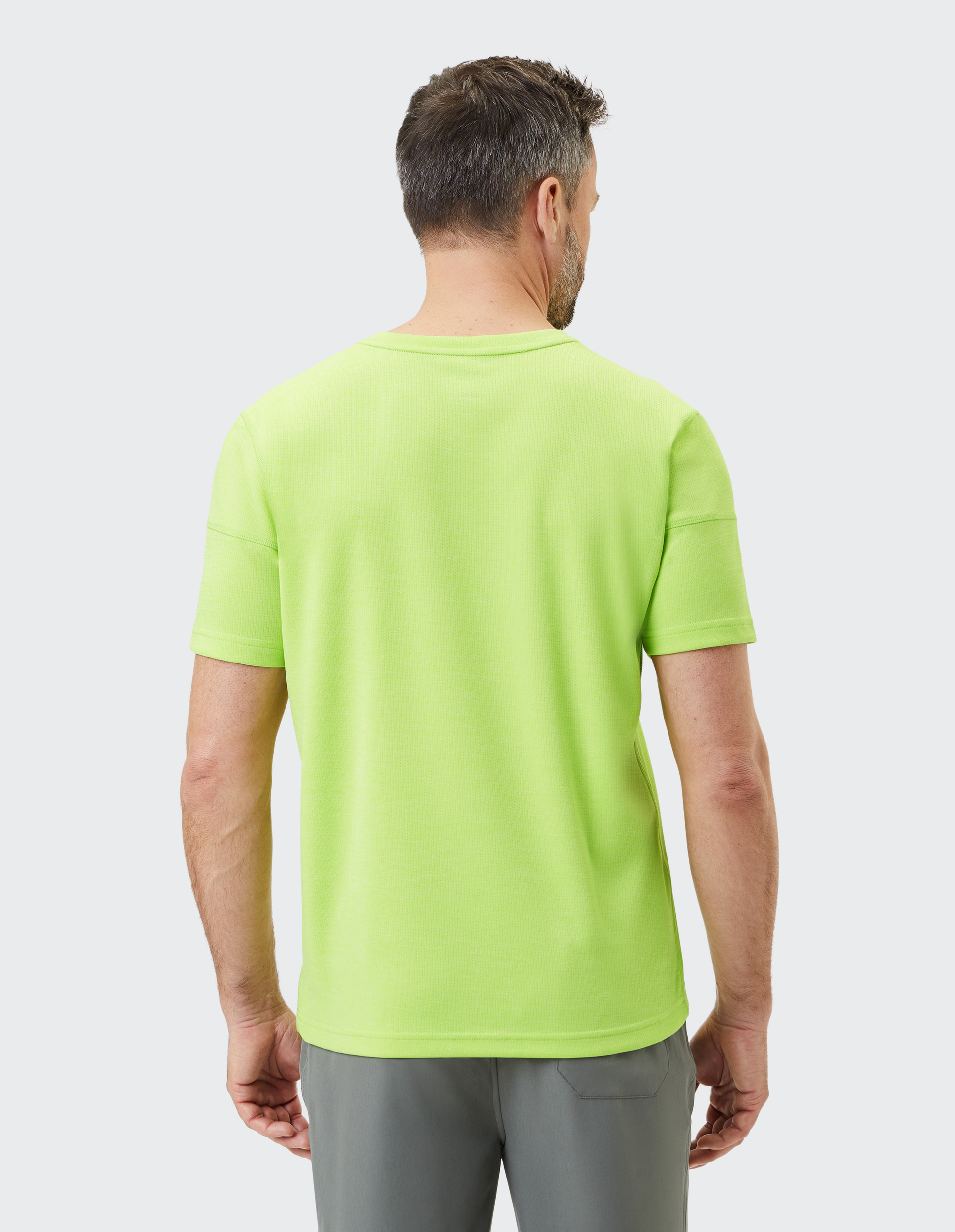 Ein Mann mit kurzen Haaren trägt ein leuchtend grünes T-Shirt und graue Hose, steht mit dem Rücken zum Betrachter vor neutralem Hintergrund.