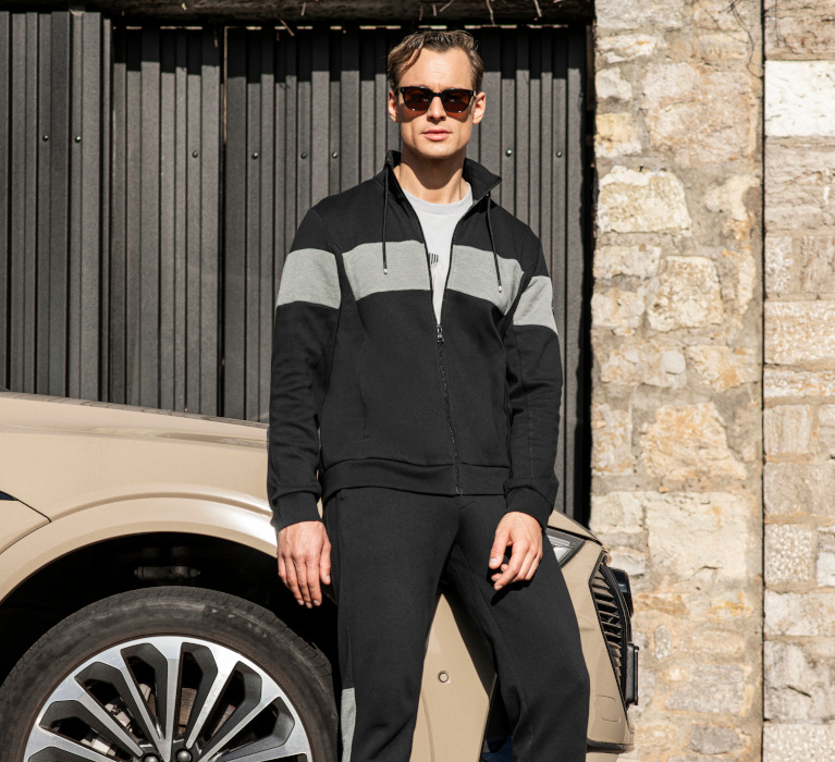 Mann steht vor einer Garage und trägt eine Sonnenbrille und eine gestreifte Jacke der Marke JOY sportswear.