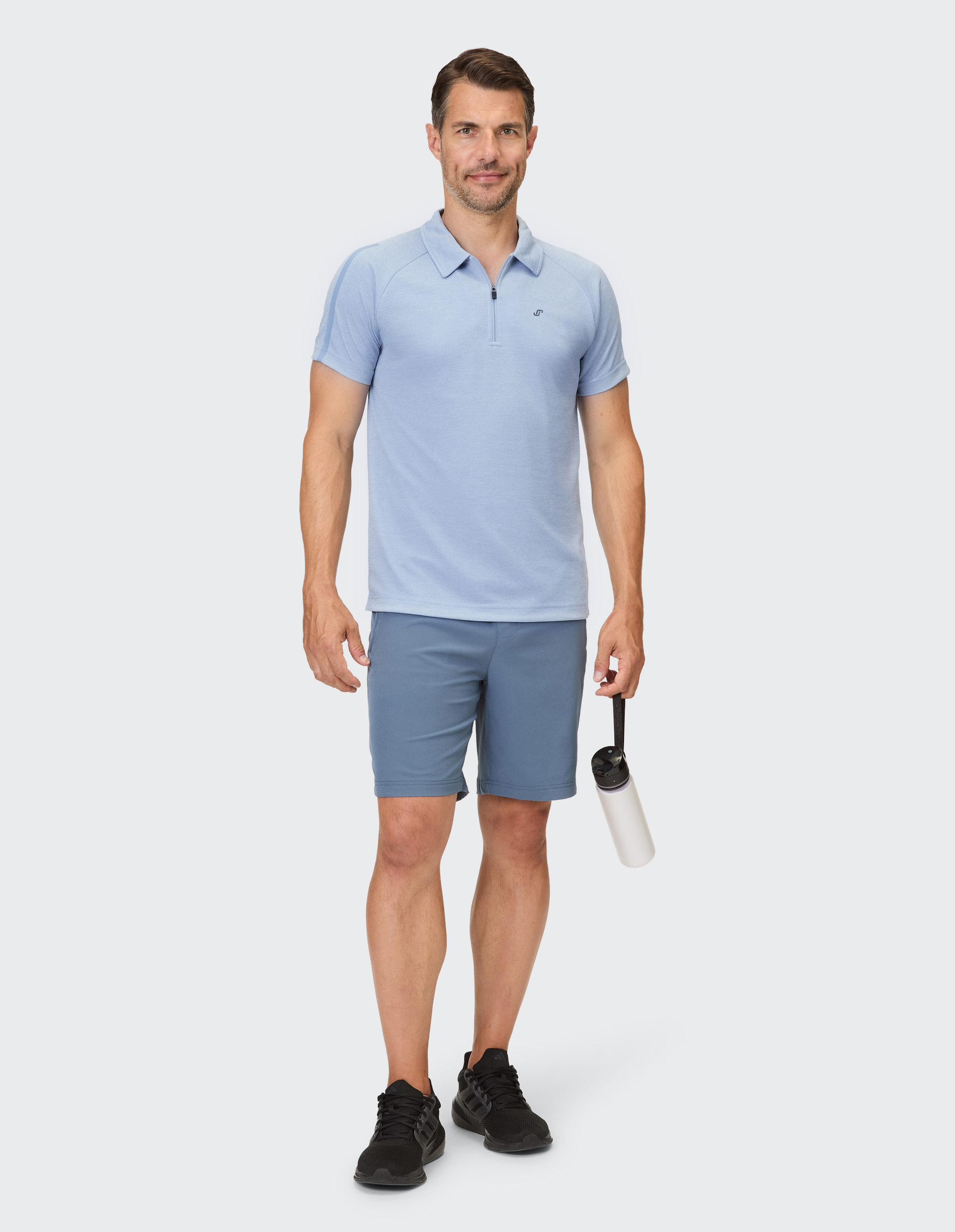 Ein Mann in einem hellblauen Poloshirt und blauen Shorts hält eine weiße Wasserflasche in der Hand.