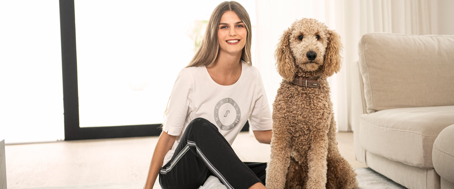 Junge Frau in heller Kleidung sitzt lächelnd auf dem Boden neben einem beigefarbenen Pudel mit Halsband.