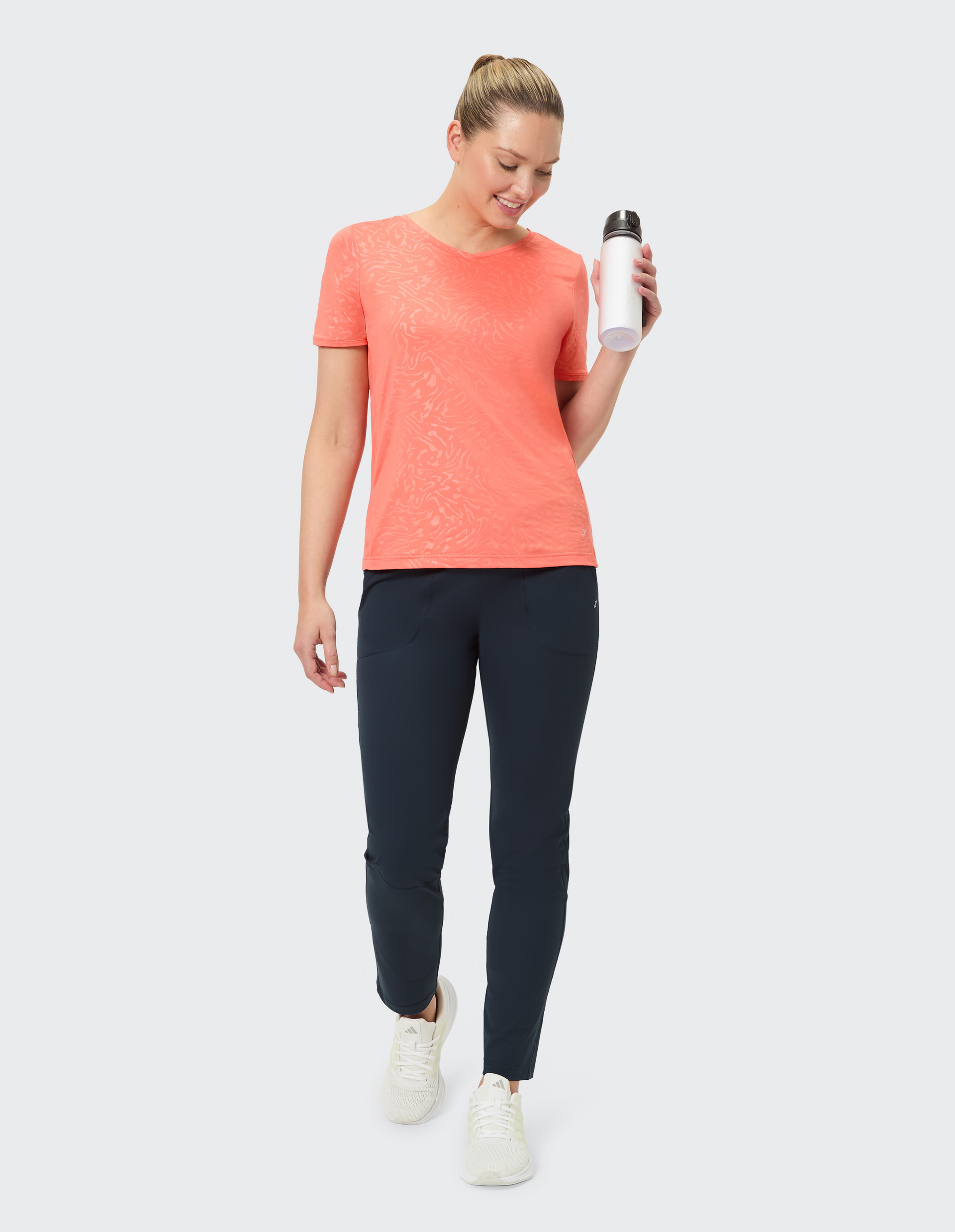 Eine Person trägt ein korallenfarbenes T-Shirt und dunkelblaue Hosen, ergänzt durch helle Sportschuhe, auf grauem Hintergrund.