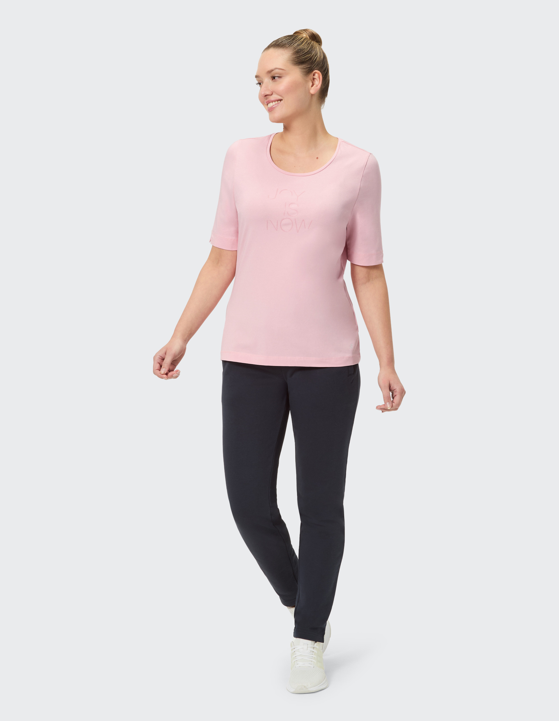 Frau in rosa T-Shirt mit Aufdruck "JOY IS NOW", dunkler Hose und weißen Turnschuhen, stehend vor einem neutralen Hintergrund.