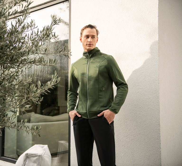 Mann in grüner Jacke der Marke JOY sportswear steht mit den Händen in den Hosentaschen neben einem Olivenbaum.