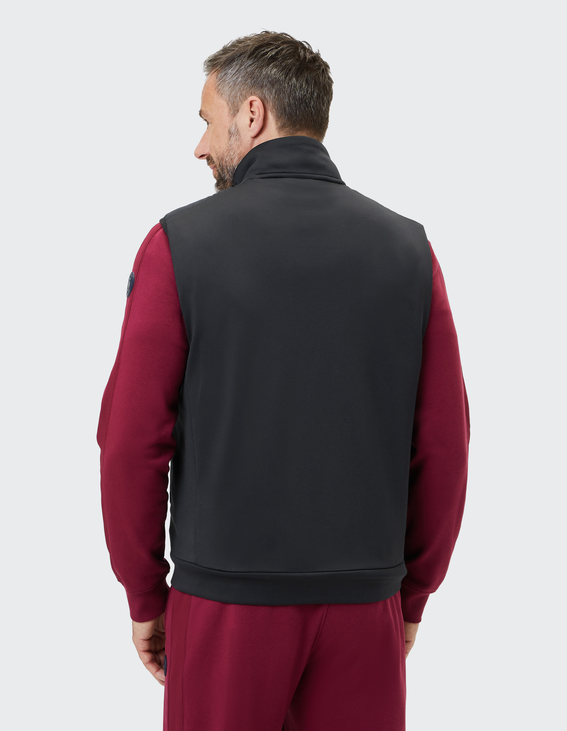 Mann von hinten in schwarzer Weste über bordeauxrotem Freizeitanzug vor weißem Hintergrund.