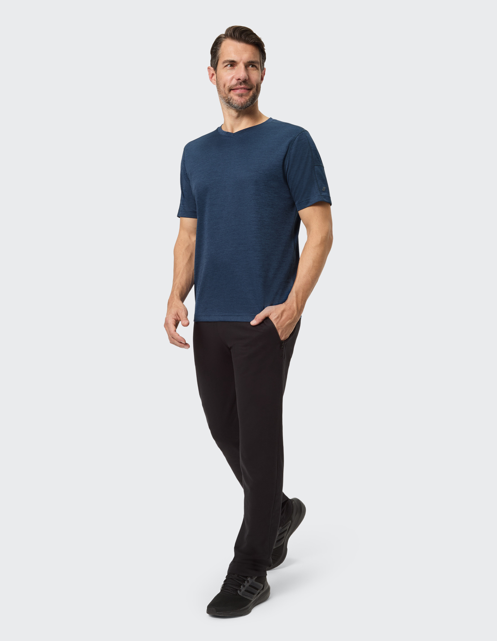 Ein lächelnder Mann trägt ein dunkelblaues T-Shirt und schwarze Hosen, stehend vor einem weißen Hintergrund.