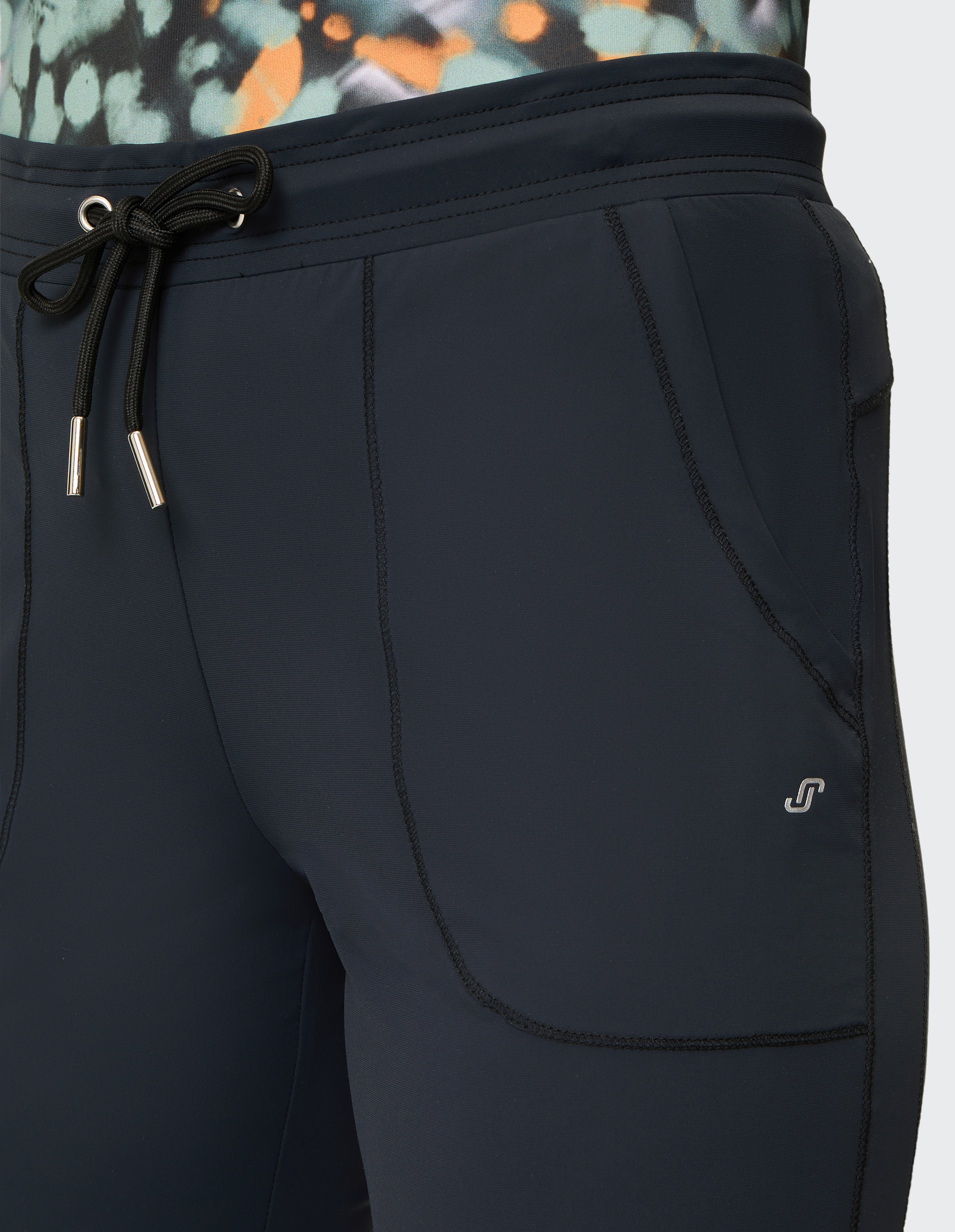 Dunkelblaue Hose mit Kordelzug, detailreicher Naht und Logo am Bein, getragen mit einem gemusterten Oberteil in Orange und Grün.