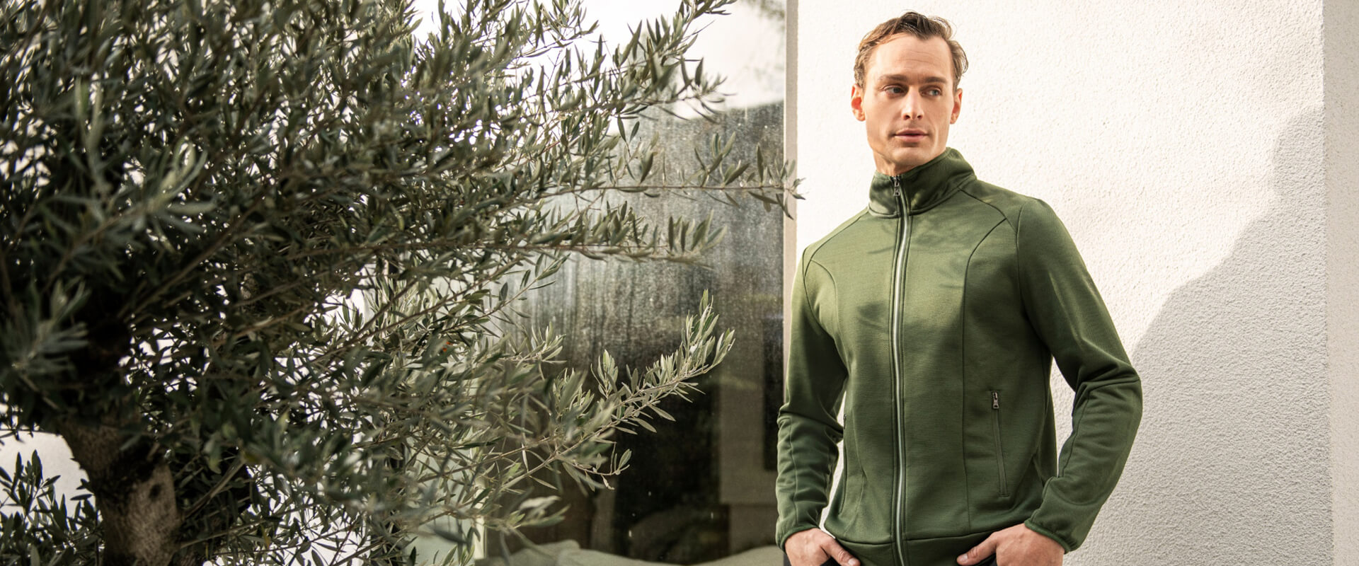 Grüner Herren-Outdoor-Fleecepullover mit durchgehendem Reißverschluss, getragen von einem Mann vor heller Hauswand neben Olivenbaum im Sonnenlicht.