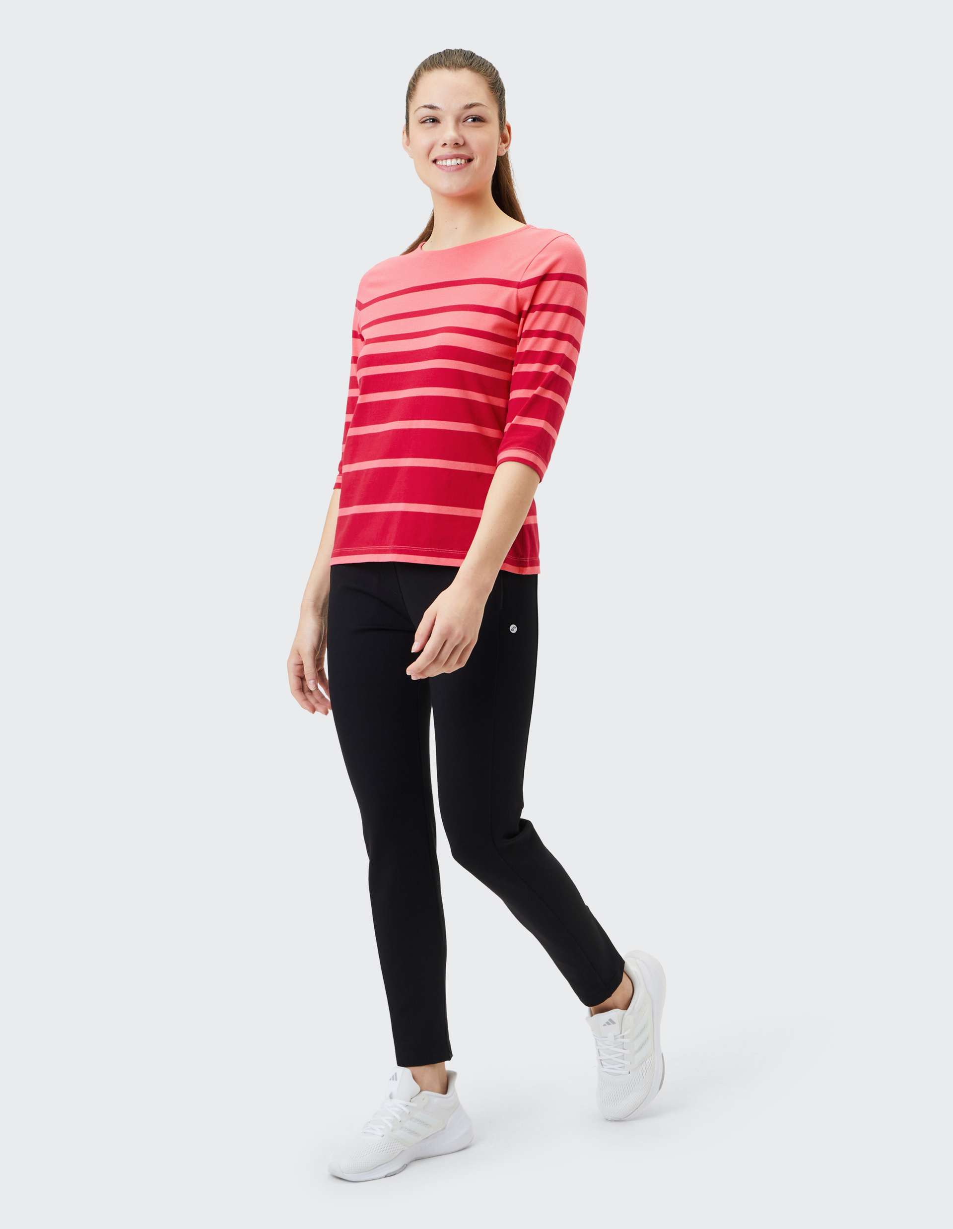 Frau in rot-gestreiftem Shirt und schwarzer Hose lächelt und geht, trägt weiße Sneakers vor neutralem Hintergrund.