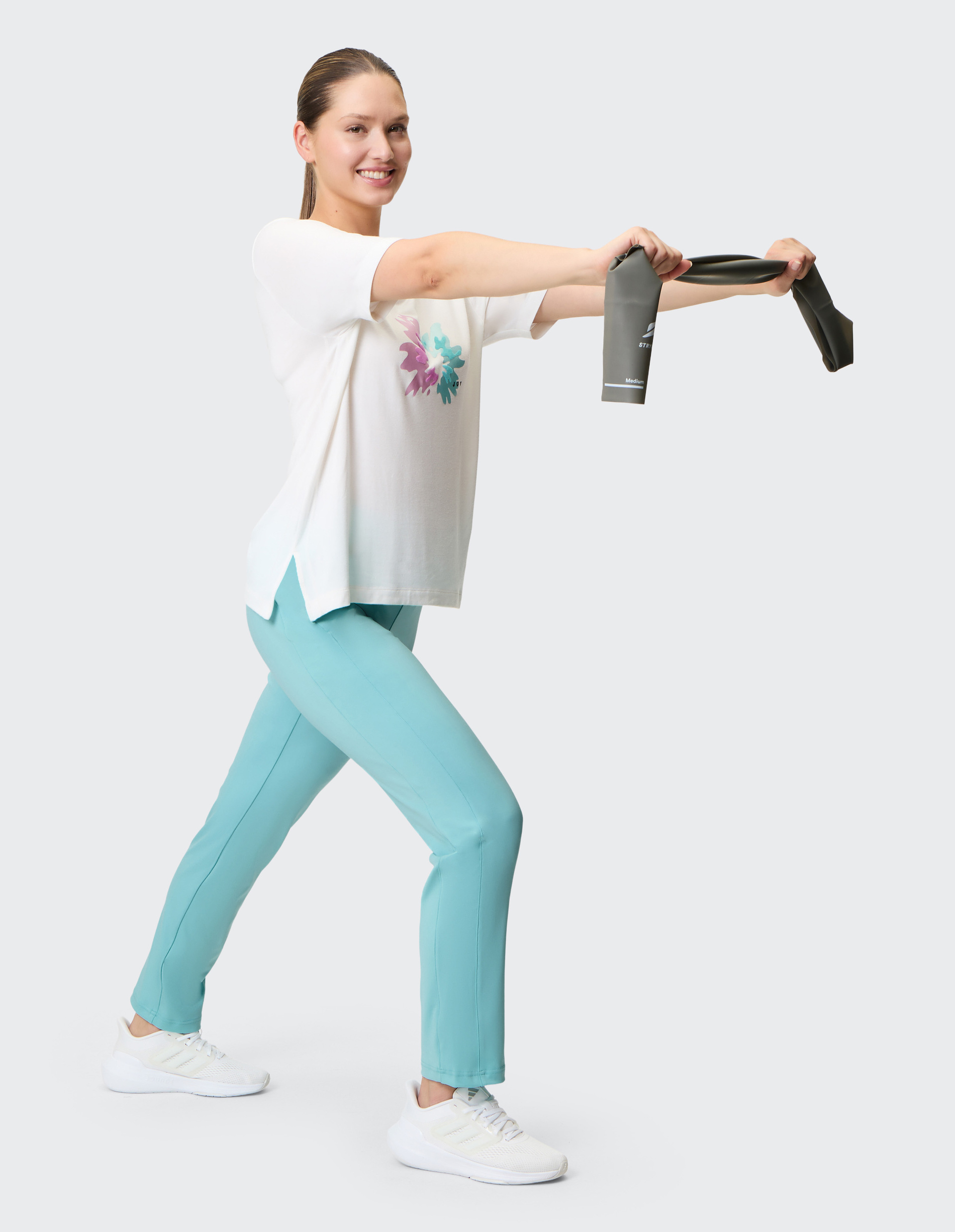 Frau in sportlicher Pose mit Widerstandsband, trägt weißes T-Shirt und türkise Hose, lächelnd, auf hellgrauem Hintergrund.