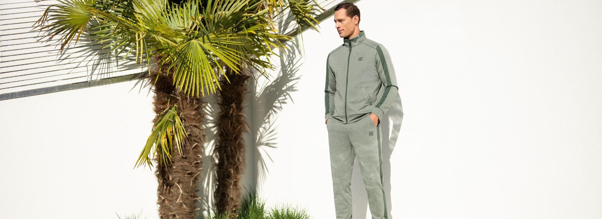 Mann in grünlichen Jogginganzug der Marke JOY sportswear steht mit den Händen in den Hosentaschen vor einem Pool.