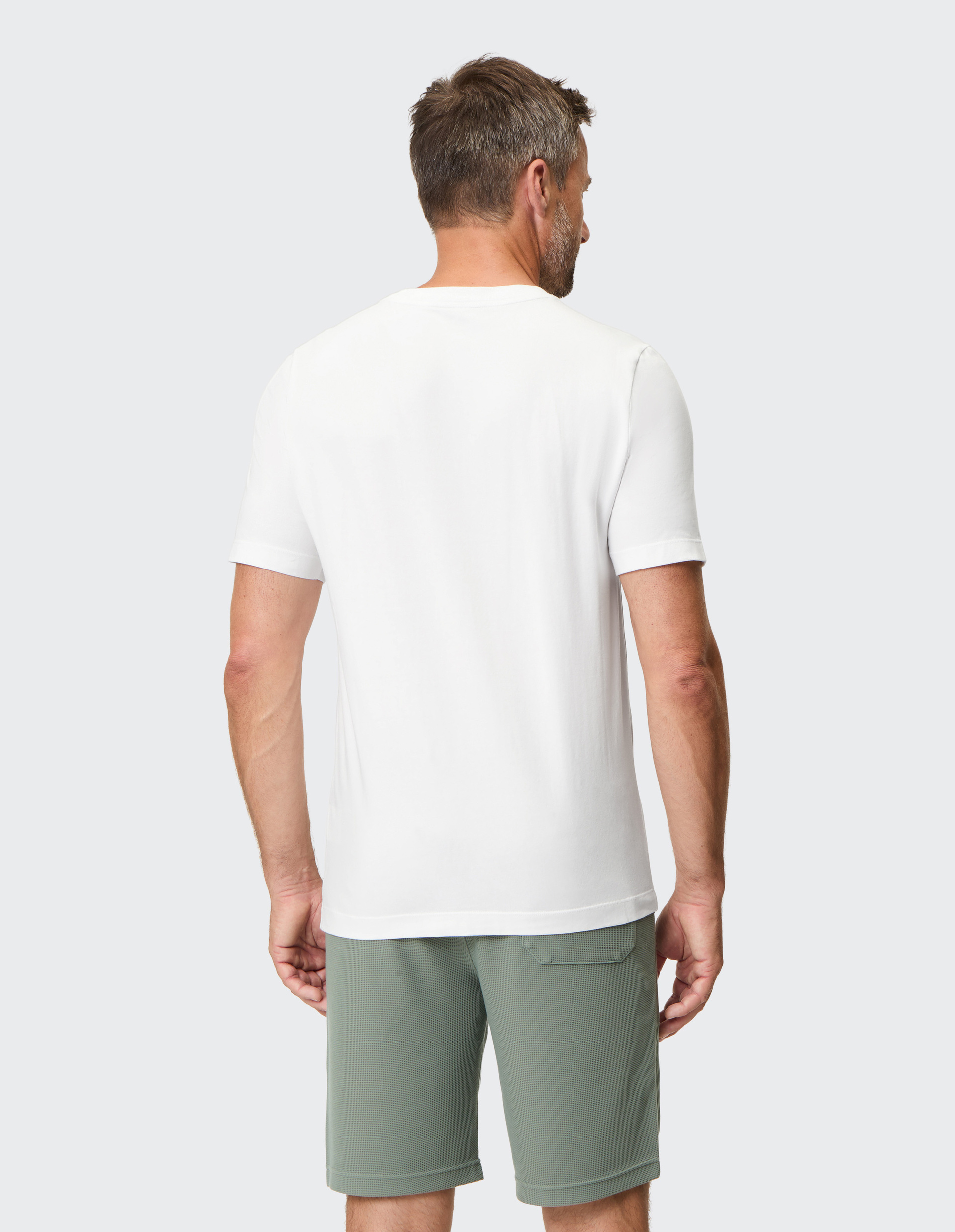 Ein Mann mit kurzem Haar trägt ein weißes T-Shirt und olivgrüne Shorts, steht mit dem Rücken zur Kamera vor grauem Hintergrund.