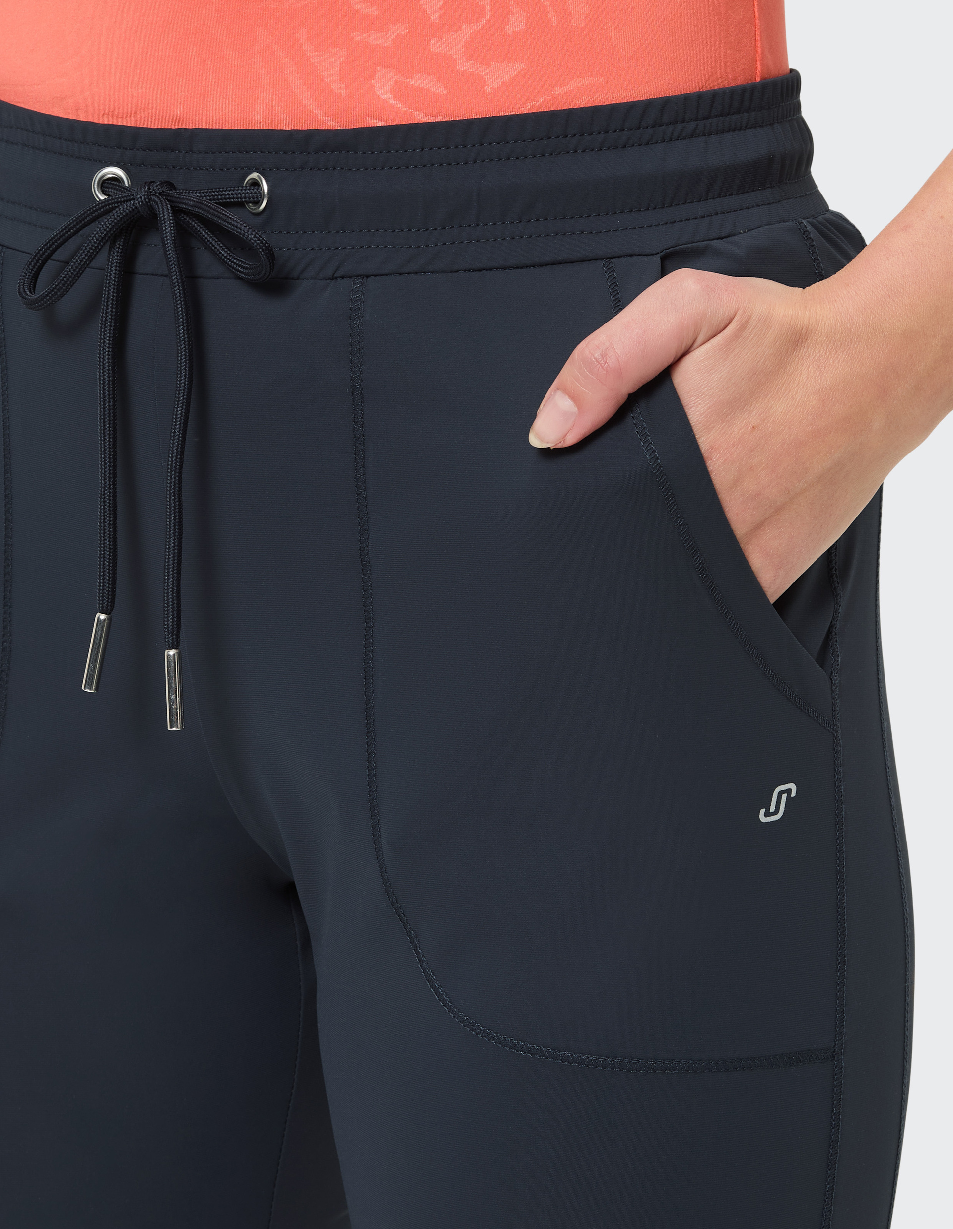 Nahaufnahme einer Person, die eine schwarze Sporthose mit Kordelzug trägt, eine Hand in der Hosentasche, auf orangem Oberteil.