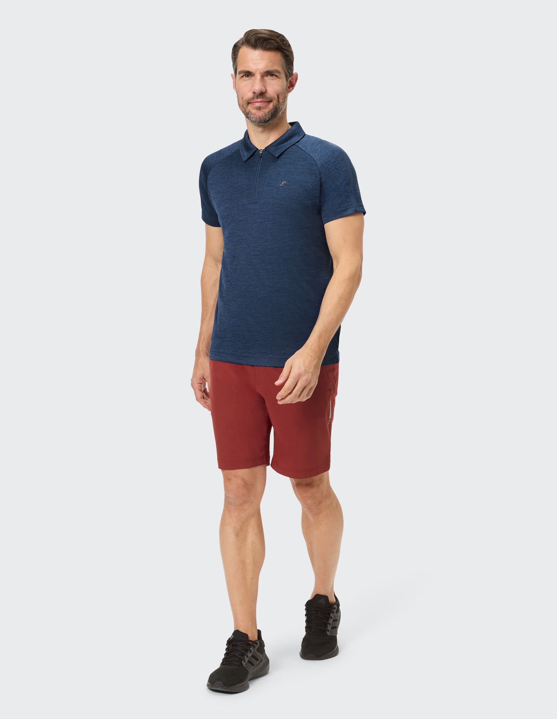 Mann in blauem Polohemd, roten Shorts und schwarzen Sneakers, steht lächelnd auf hellem Hintergrund.
