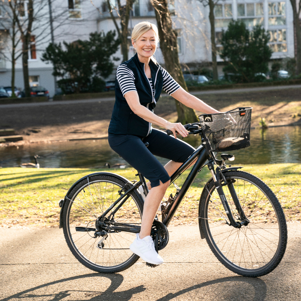 Frau fährt auf einem Fahrrad auf einem Fahrradweg vor einem Fluss und lächelt frontal in die Kamera.
