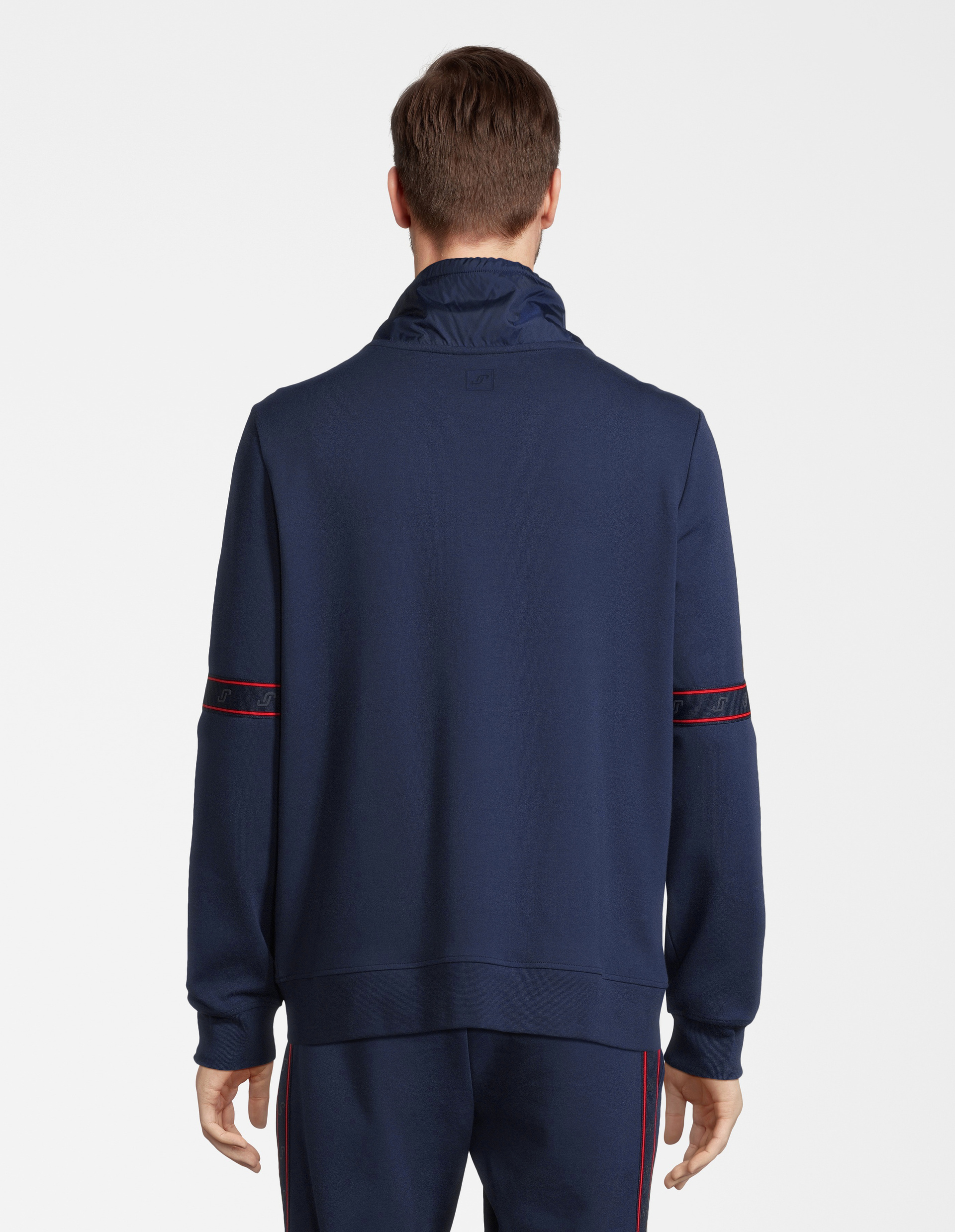 Eine Person trägt einen blauen Trainingsanzug mit roten Streifen, stehend und von hinten aufgenommen vor weißem Hintergrund.