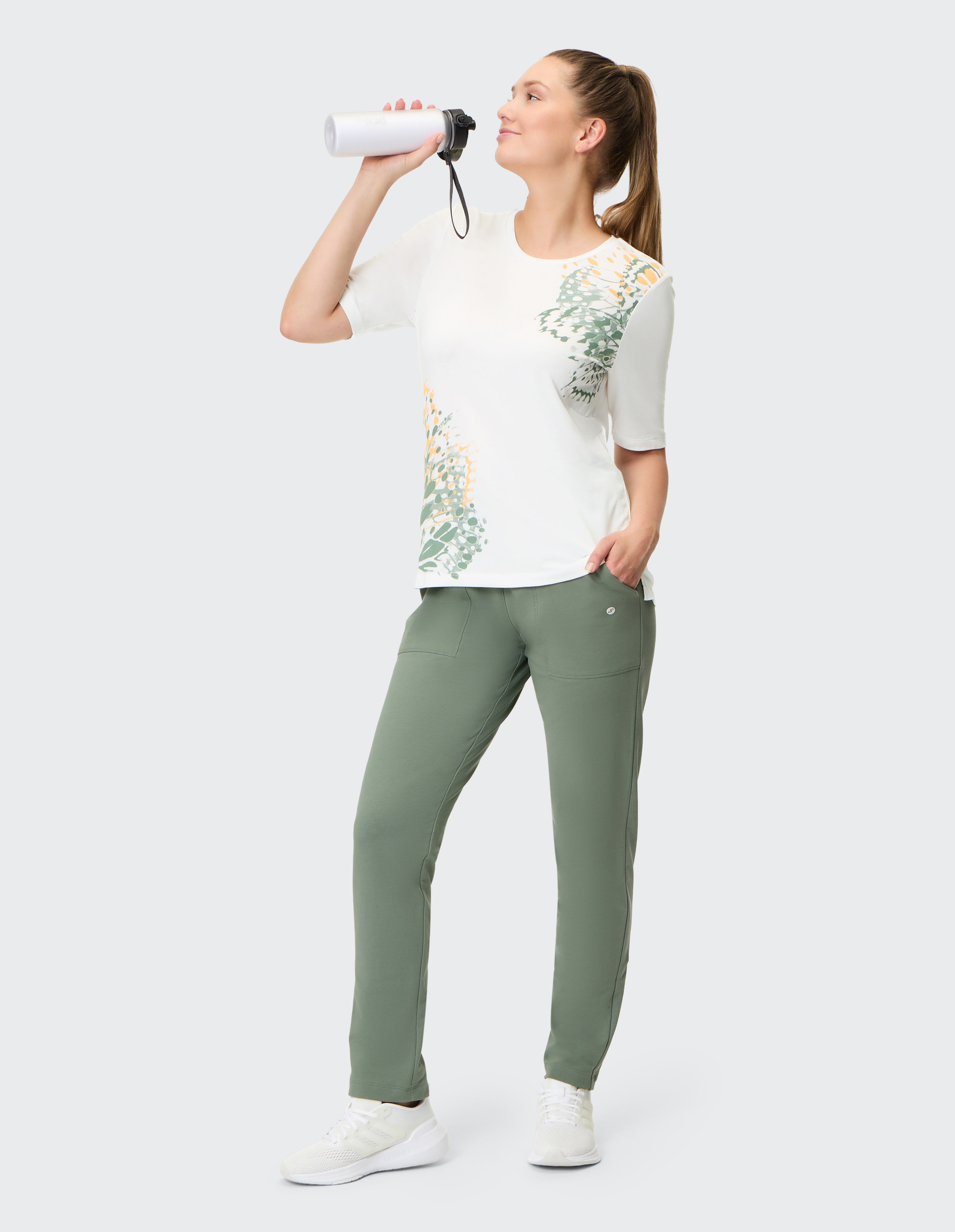Eine Frau in sportlicher Kleidung trinkt aus einer Wasserflasche, trägt ein gemustertes weißes Shirt und grüne Hosen.