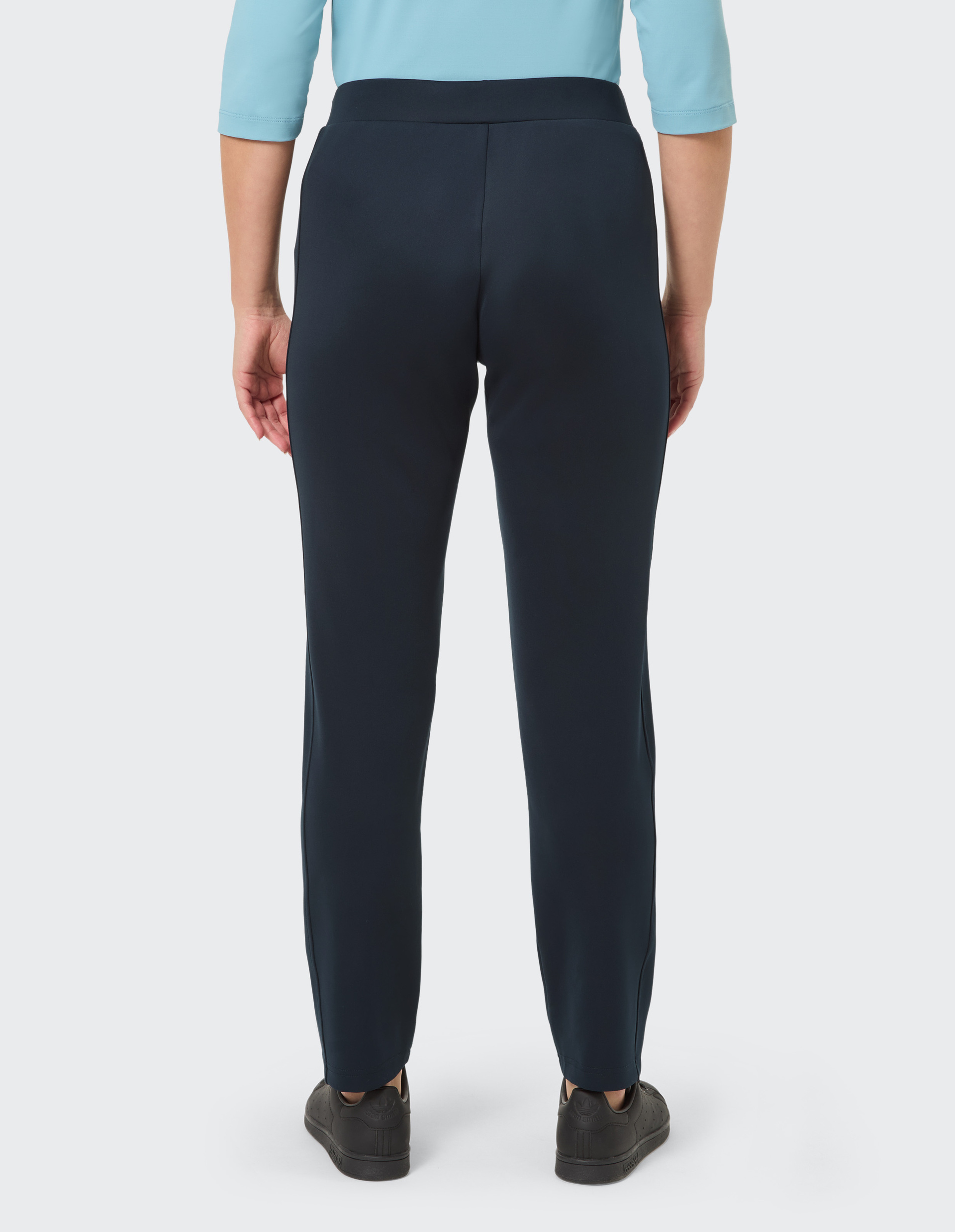 Person von hinten in schwarzer Hose und türkisfarbenem Oberteil, stehend auf hellem Hintergrund, mit schwarzen Schuhen.