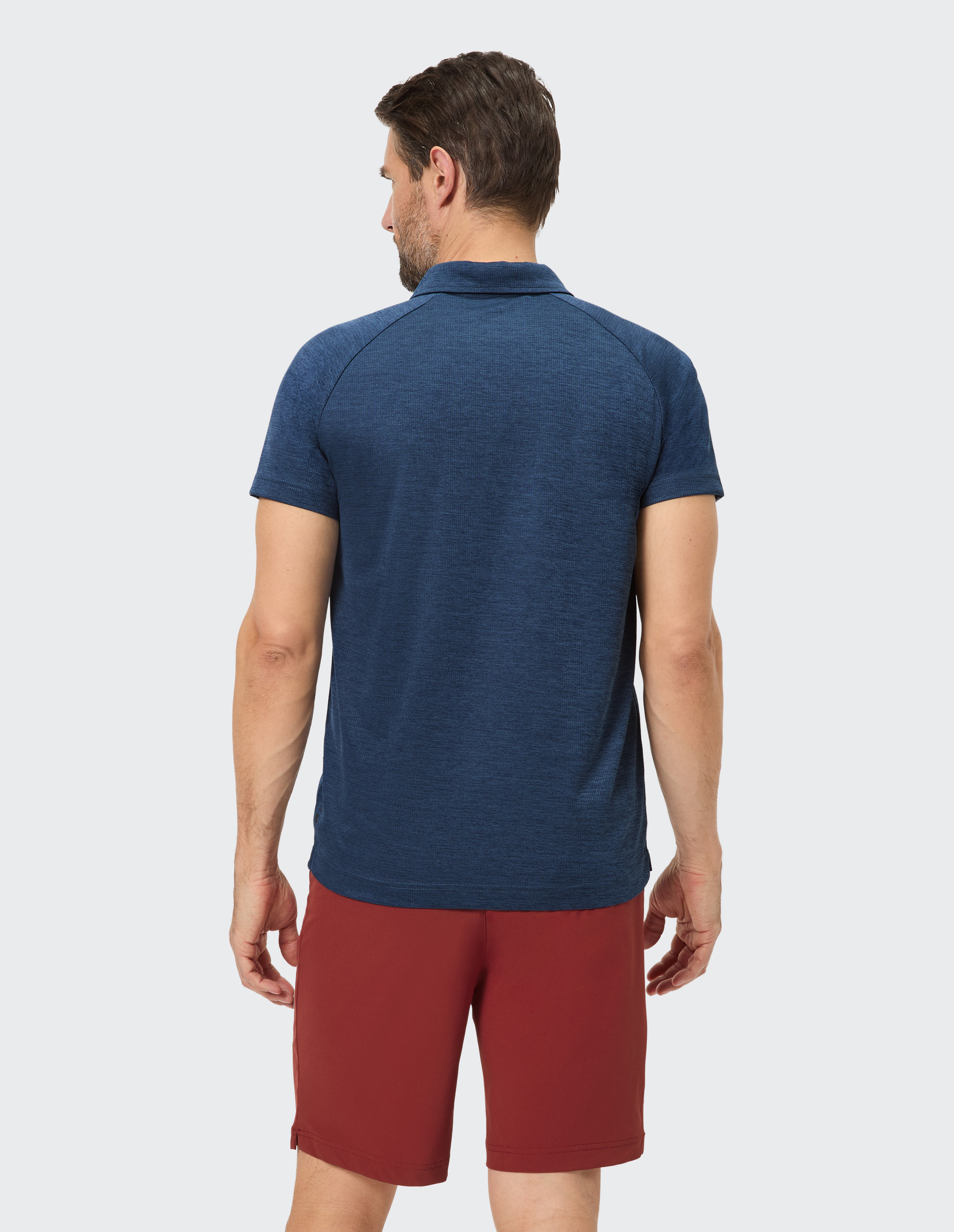 Ein Mann von hinten in einem dunkelblauen Polohemd und roten Shorts, steht vor einem neutralen Hintergrund.