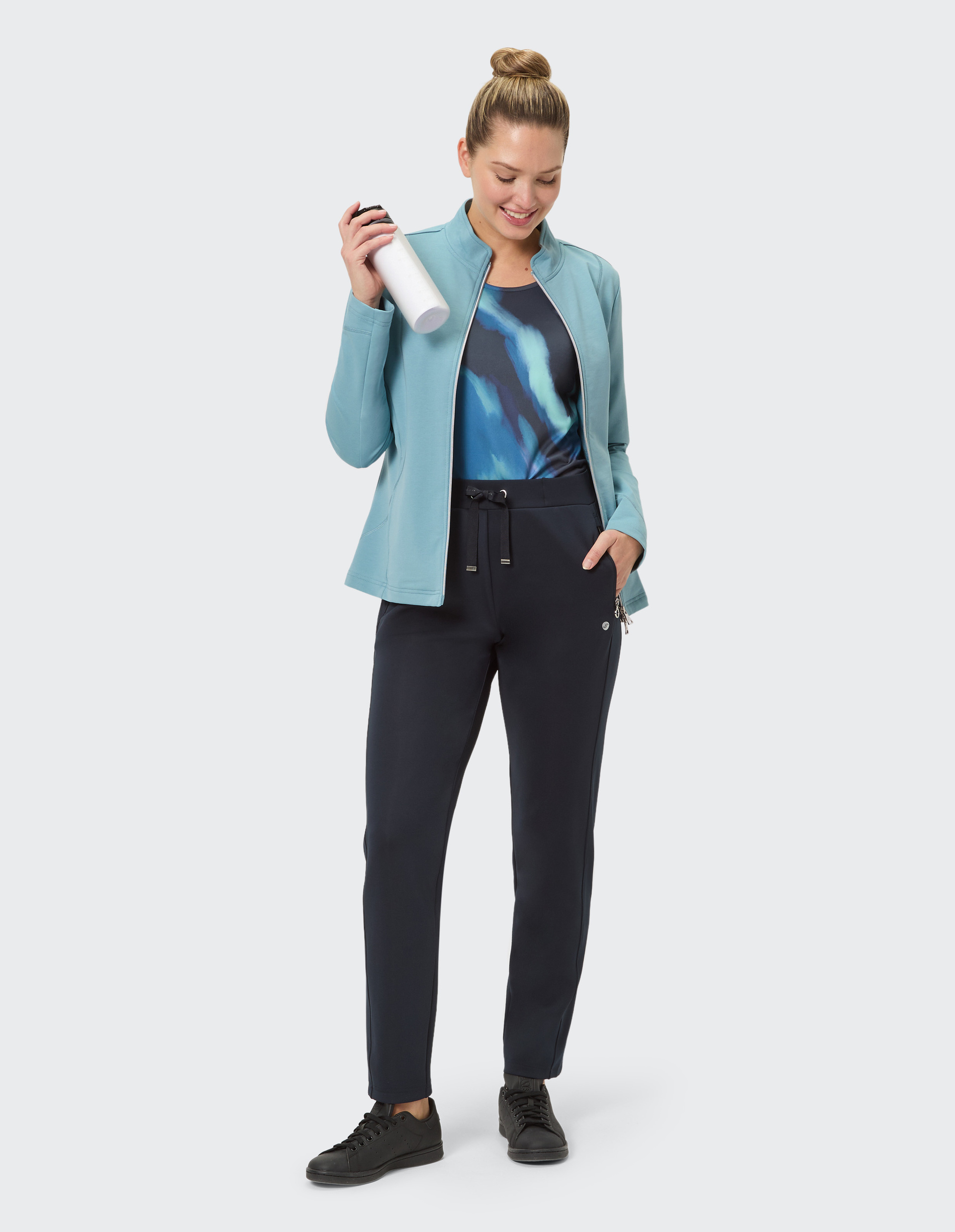 Frau in hellblauer Jacke mit Reißverschluss und dunkler Hose steht vor hellem Hintergrund, lächelt dezent, Haare hochgesteckt.