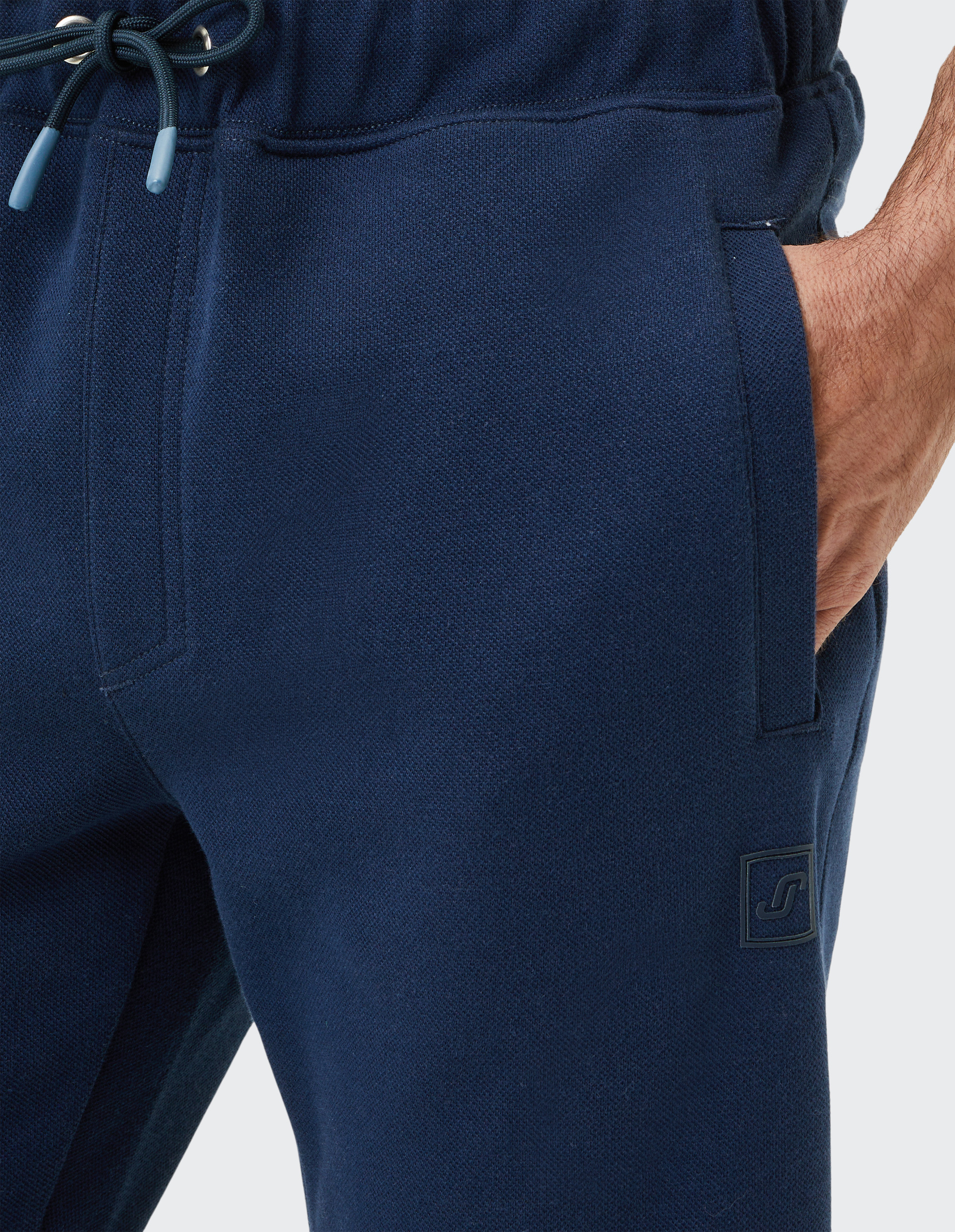 Nahaufnahme einer dunkelblauen Jogginghose mit Kordelzug und eingesteckter Hand in der Seitentasche auf grauem Hintergrund.