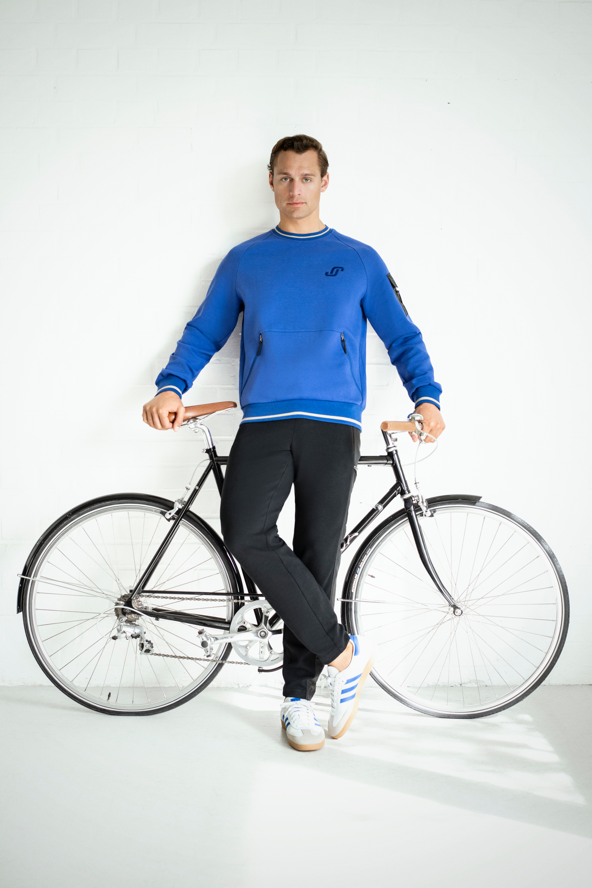 Mann in blauem Pullover und schwarzen Hosen lehnt lässig an einem schwarzen Fahrrad, trägt weiße Turnschuhe mit blauen Streifen.