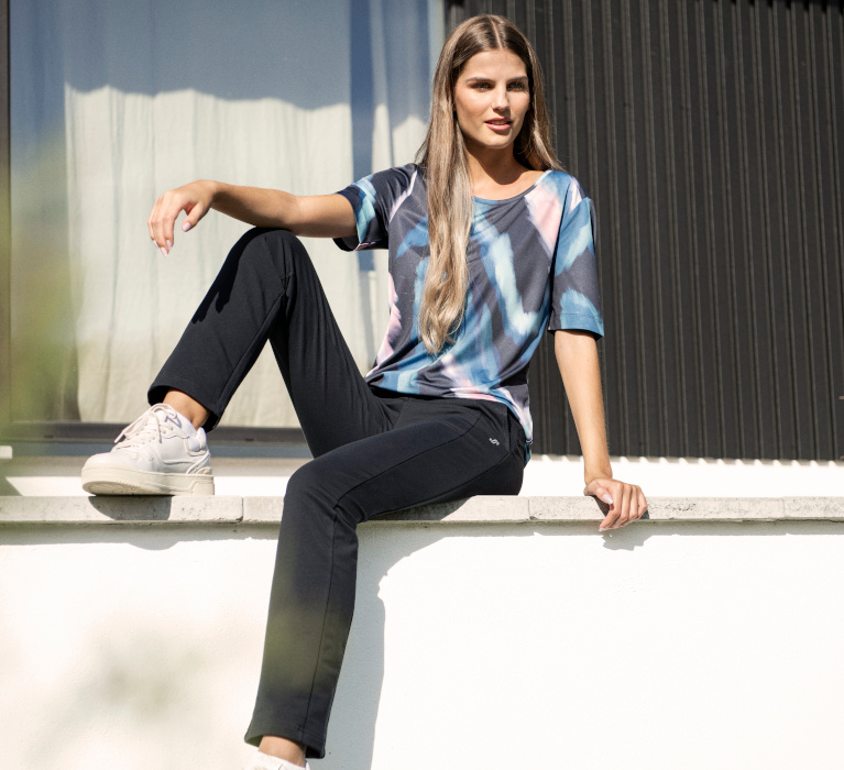 Auf einer Mauer sitzenden Frau in dunkelblauer Jogginghose und T-Shirt mit grafischem Print in den Farben hellblau, dunkelblau und rosa.