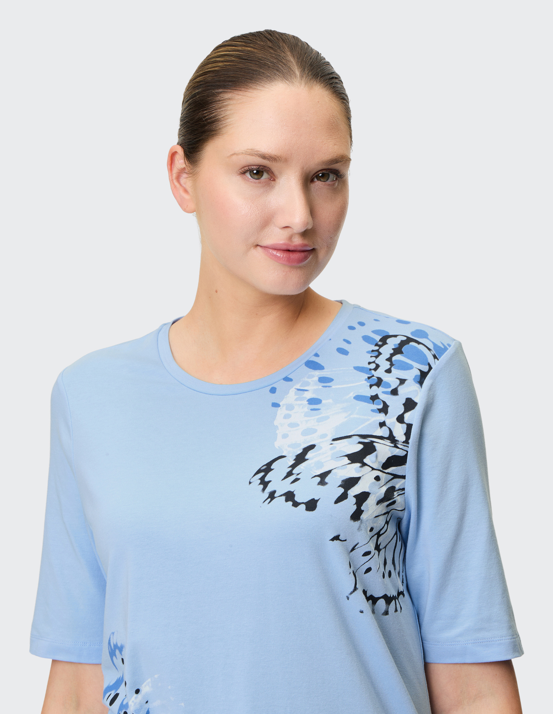 Frau mit zurückgebundenem Haar in hellblauem T-Shirt mit abstraktem schwarz-blauem Muster, vor hellem Hintergrund.