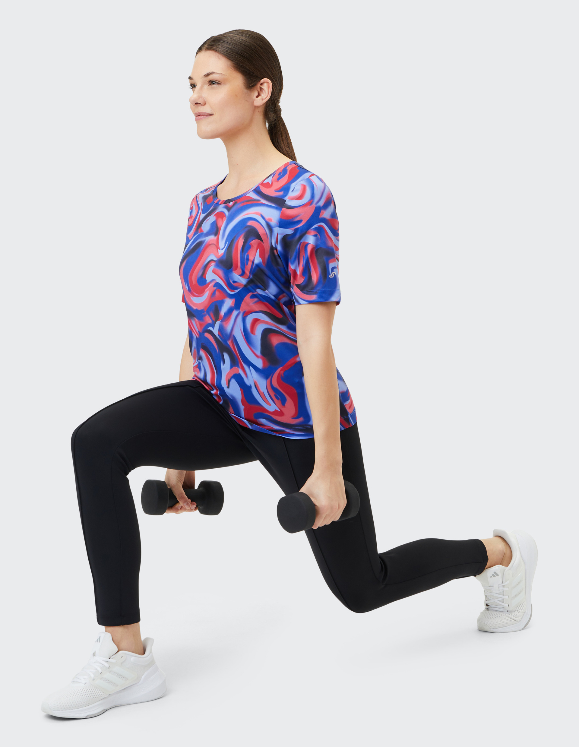 Eine Frau in bunter Sportkleidung macht einen Ausfallschritt mit Hanteln, lächelt und trägt weiße Turnschuhe auf hellem Hintergrund.