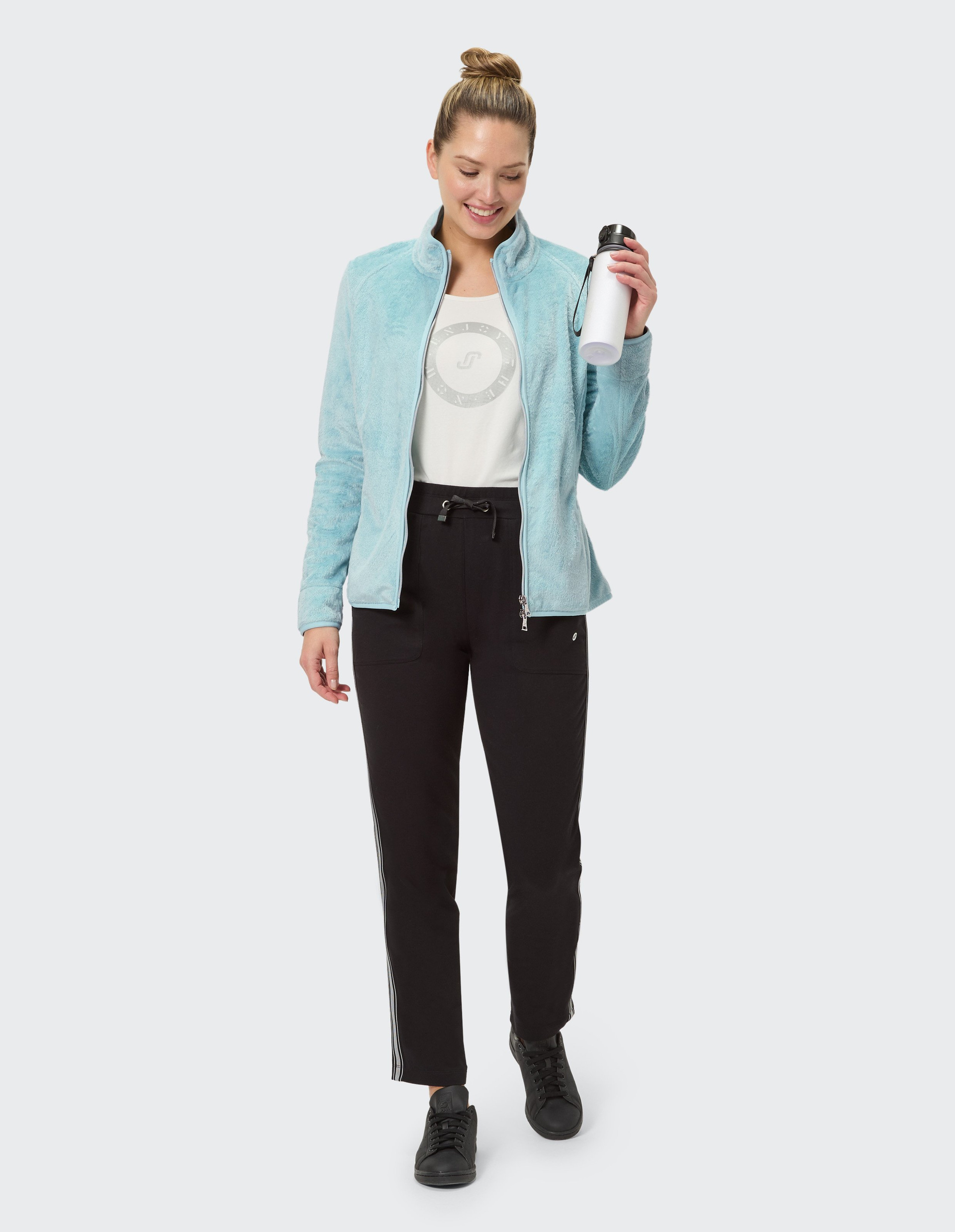 Frau in hellblauer Jacke und schwarzen Hosen steht frontal, lächelt leicht, trägt schwarze Schuhe; weißer Hintergrund.