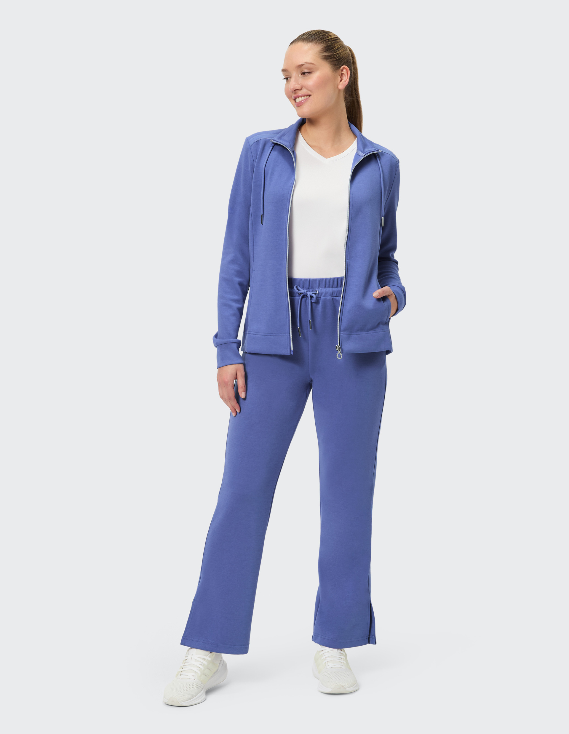 Frau in blauer Freizeitkleidung mit Reißverschlussjacke und Hose, steht aufrecht, lächelt leicht, trägt weiße Turnschuhe.