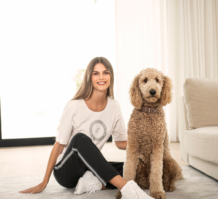 Frau in weißem T-Shirt mit Print der Marke JOY sportswear auf dem Boden sitzend und den Hund neben sich streichelnd.