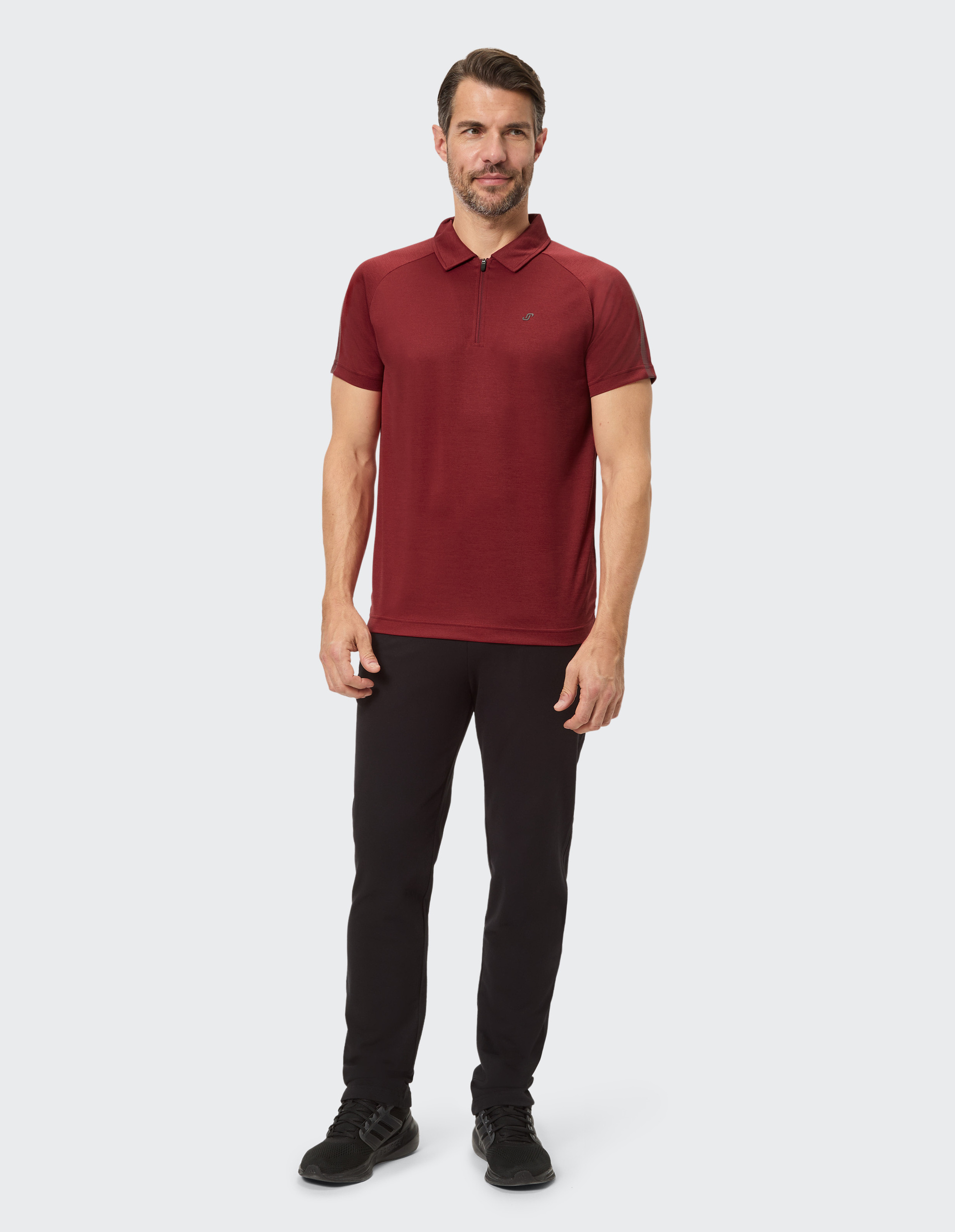 Ein Mann in rotem Polohemd und schwarzer Jogginghose hält eine weiße Wasserflasche, stehend vor neutralem Hintergrund.