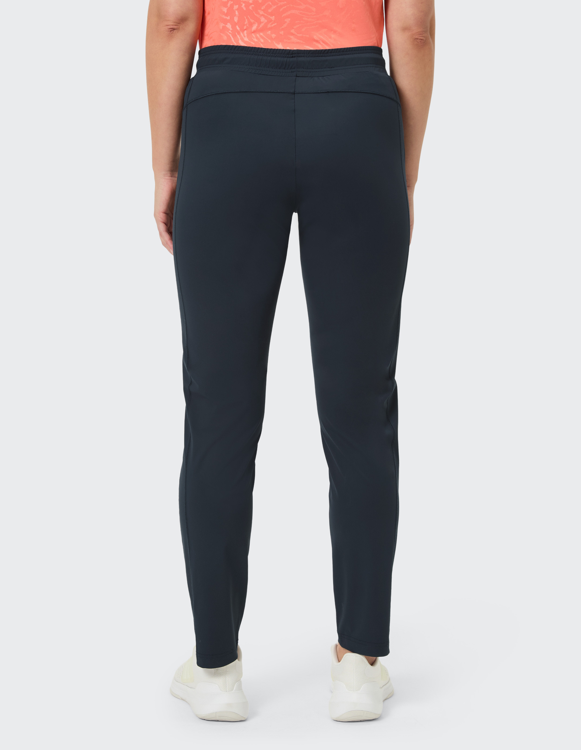 Rückansicht einer Person in dunkelblauer Sporthose, orangefarbenem Top und weißen Sneakern vor neutralem Hintergrund.