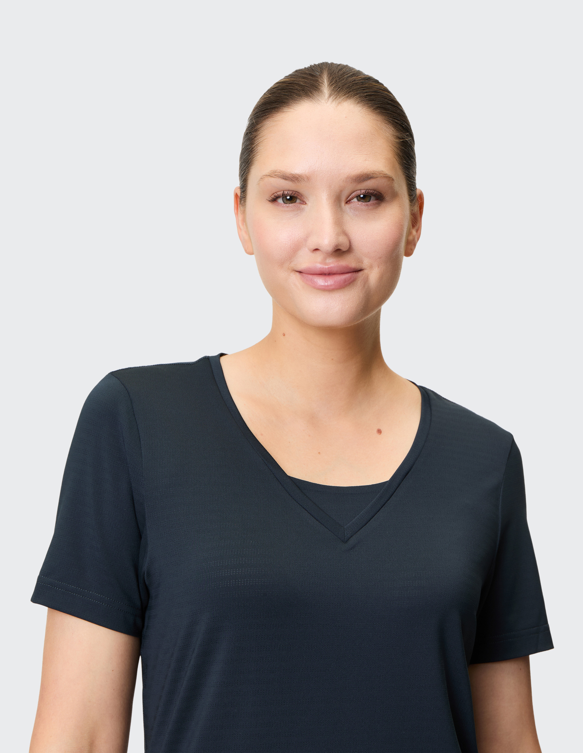 Frau mit dunklem T-Shirt und gekämmtem Haar lächelt leicht vor neutralem Hintergrund.