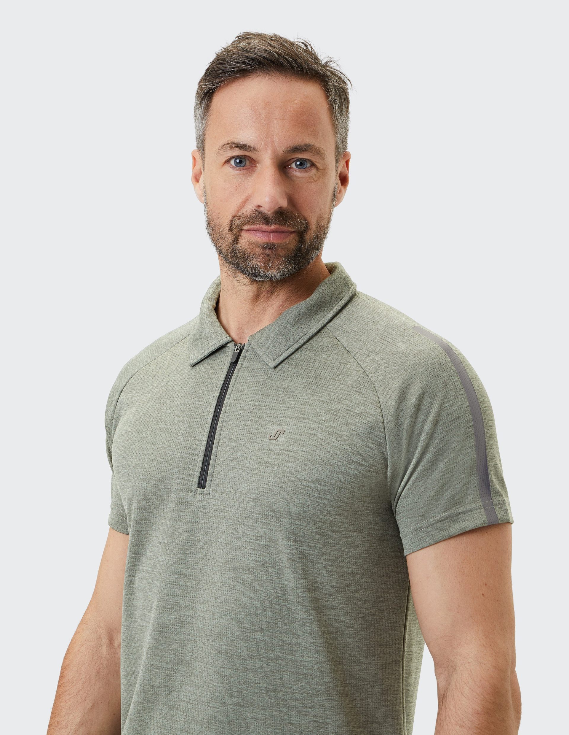 Ein Mann mit kurzem Haar trägt ein graugrünes Polohemd mit Reißverschluss und steht vor einem hellen Hintergrund.