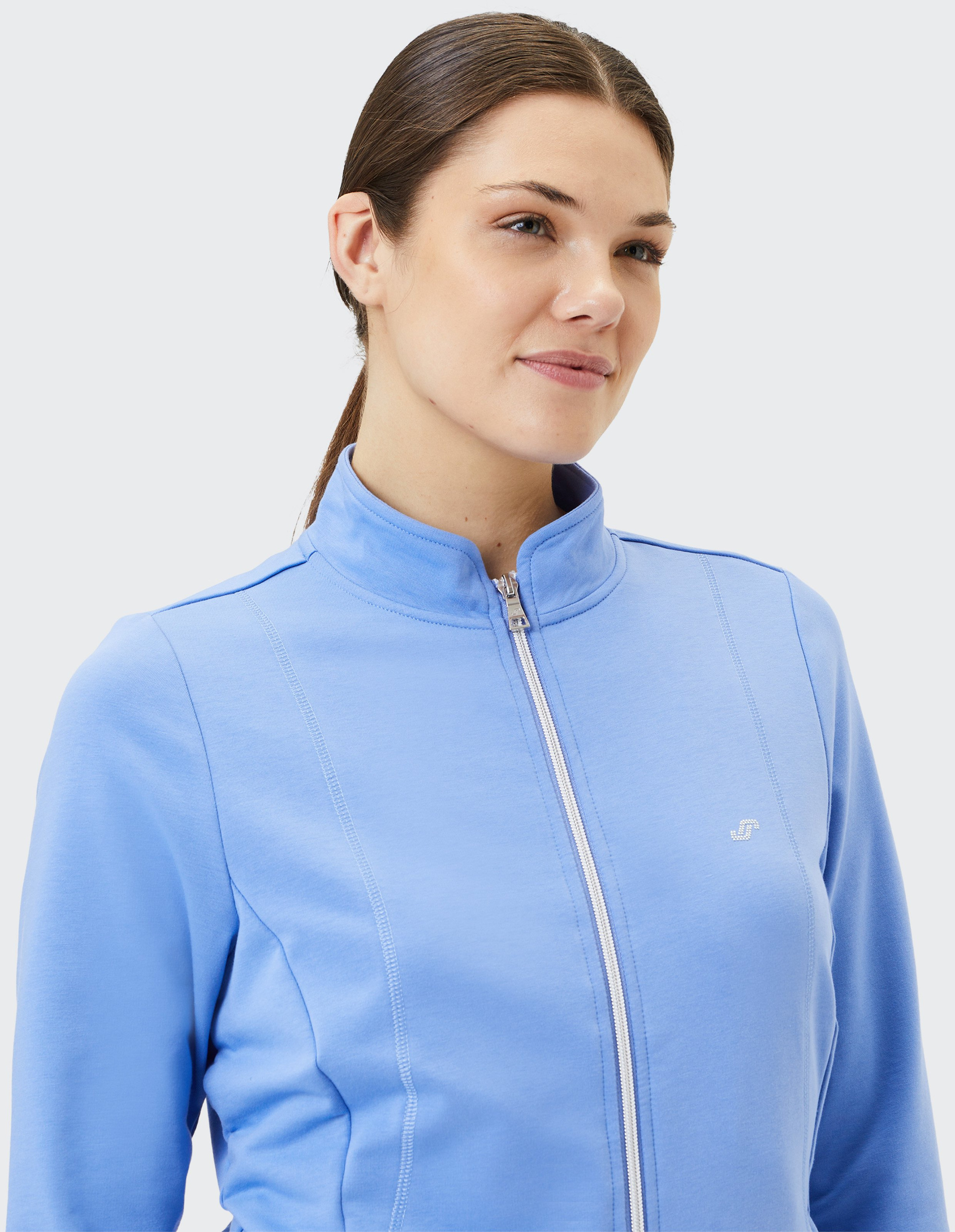 Eine Frau mit zurückgebundenem Haar trägt eine hellblaue Jacke mit Reißverschluss und kleiner Logostickerei auf der Brust.