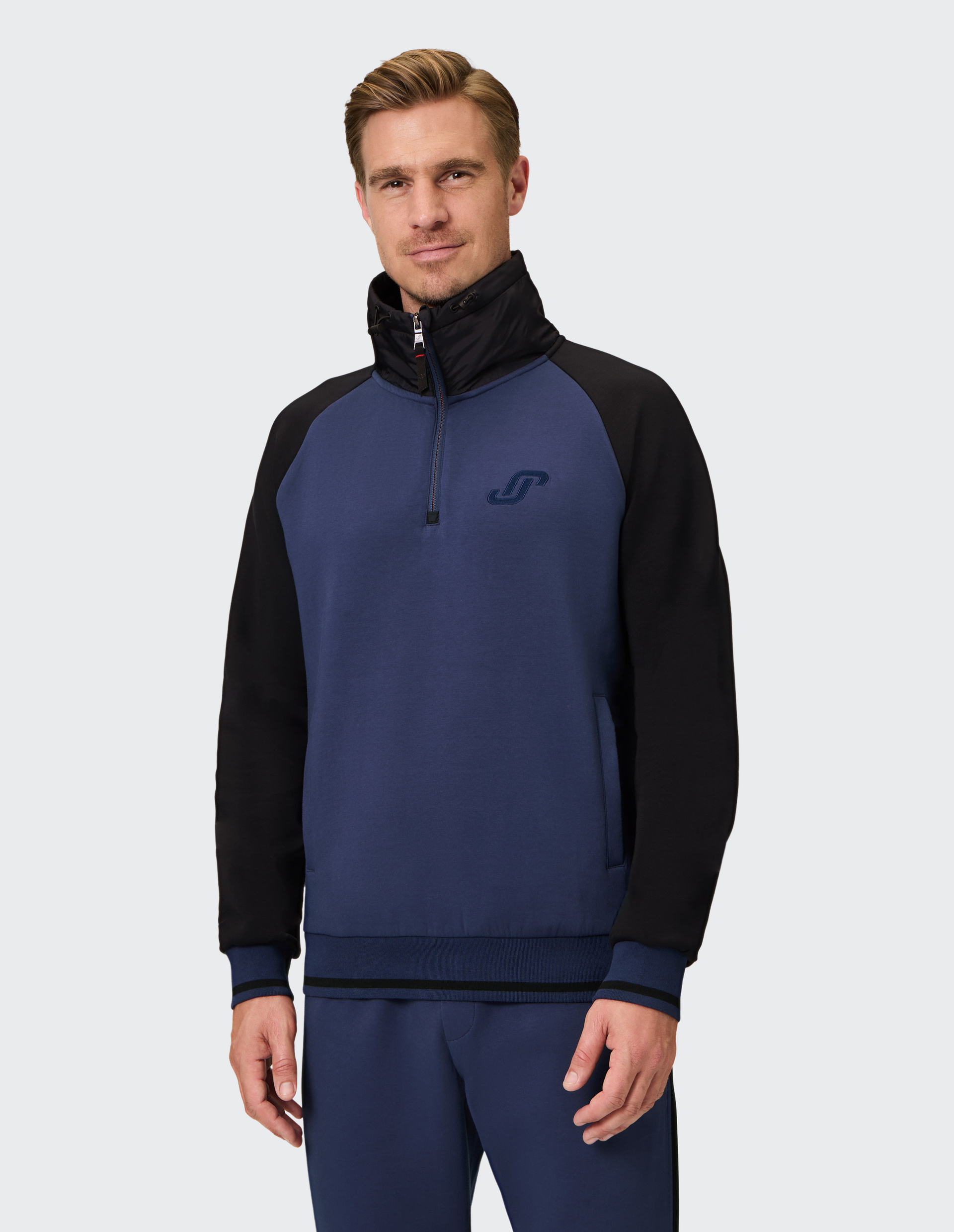 Blauer Pullover mit schwarzem Kragen und Ärmeln, Halbreißverschluss, hohem Kragen und dezentem Logo vorne auf der Brust.