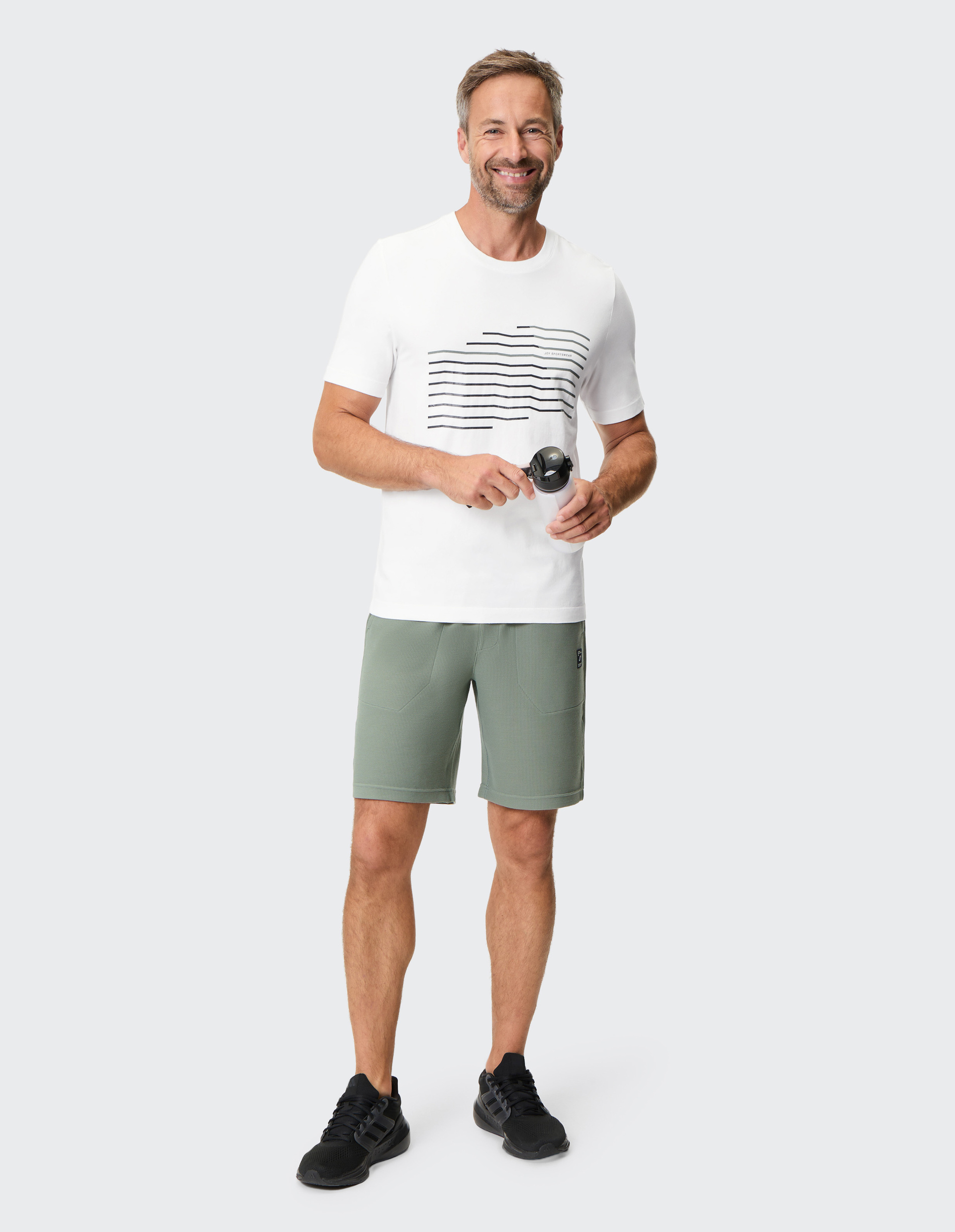 Ein lächelnder Mann trägt ein weißes T-Shirt mit Streifen, grüne Shorts und schwarze Turnschuhe, hält eine Wasserflasche in der Hand.