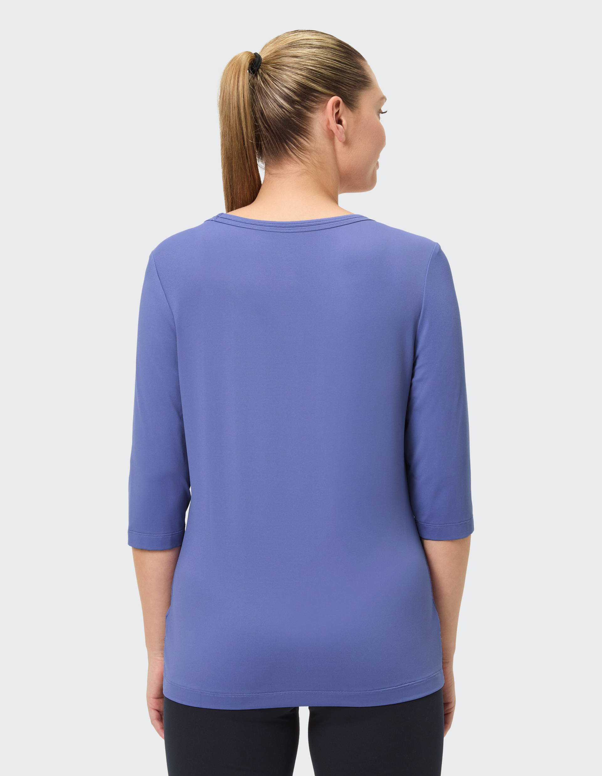 Frau mit Pferdeschwanz trägt ein blaues, dreiviertelärmliges Shirt und schwarze Hose, Rückenansicht vor grauem Hintergrund.