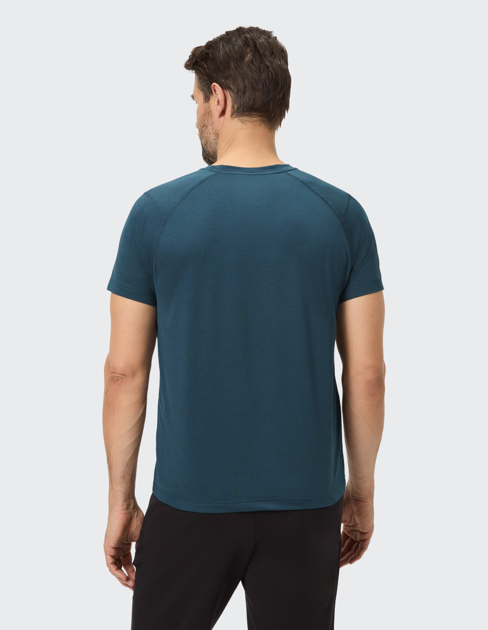 Ein Mann mit braunen Haaren trägt ein dunkelblaues T-Shirt und schwarze Hose, von hinten betrachtet, auf weißem Hintergrund.