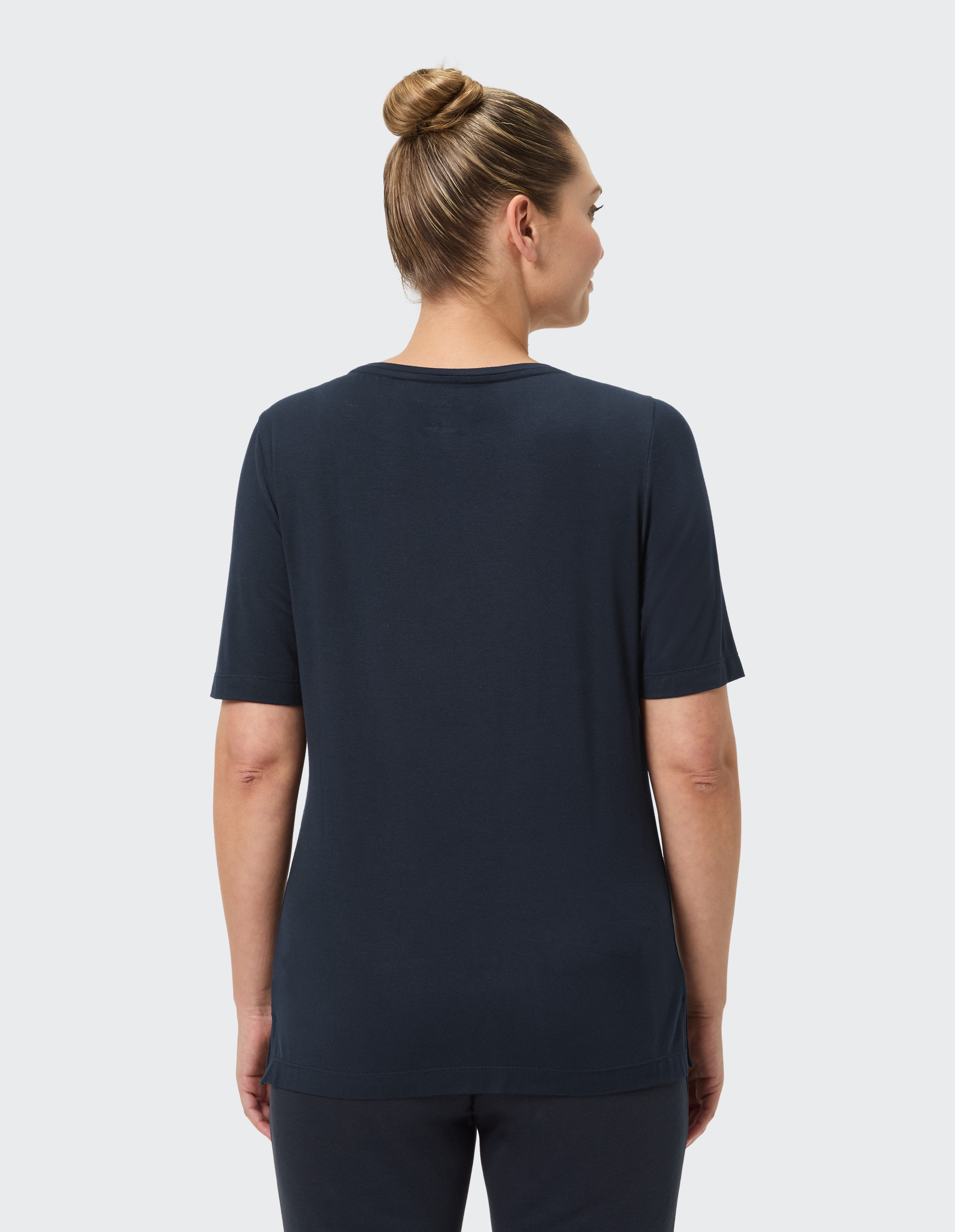 Eine Person mit Dutt trägt ein dunkelblaues T-Shirt und sieht von hinten nach rechts, der Hintergrund ist hellgrau.