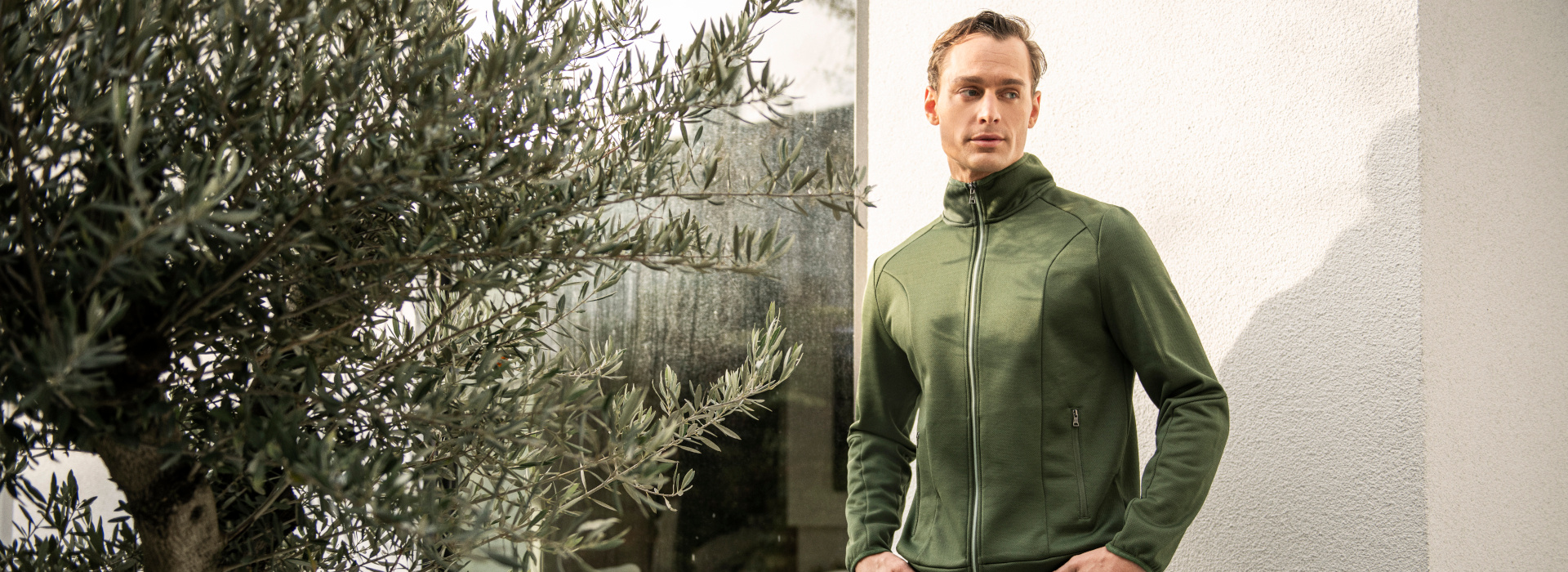 Mann in grüner Jacke der Marke JOY sportswear steht mit den Händen in den Hosentaschen neben einem Olivenbaum.