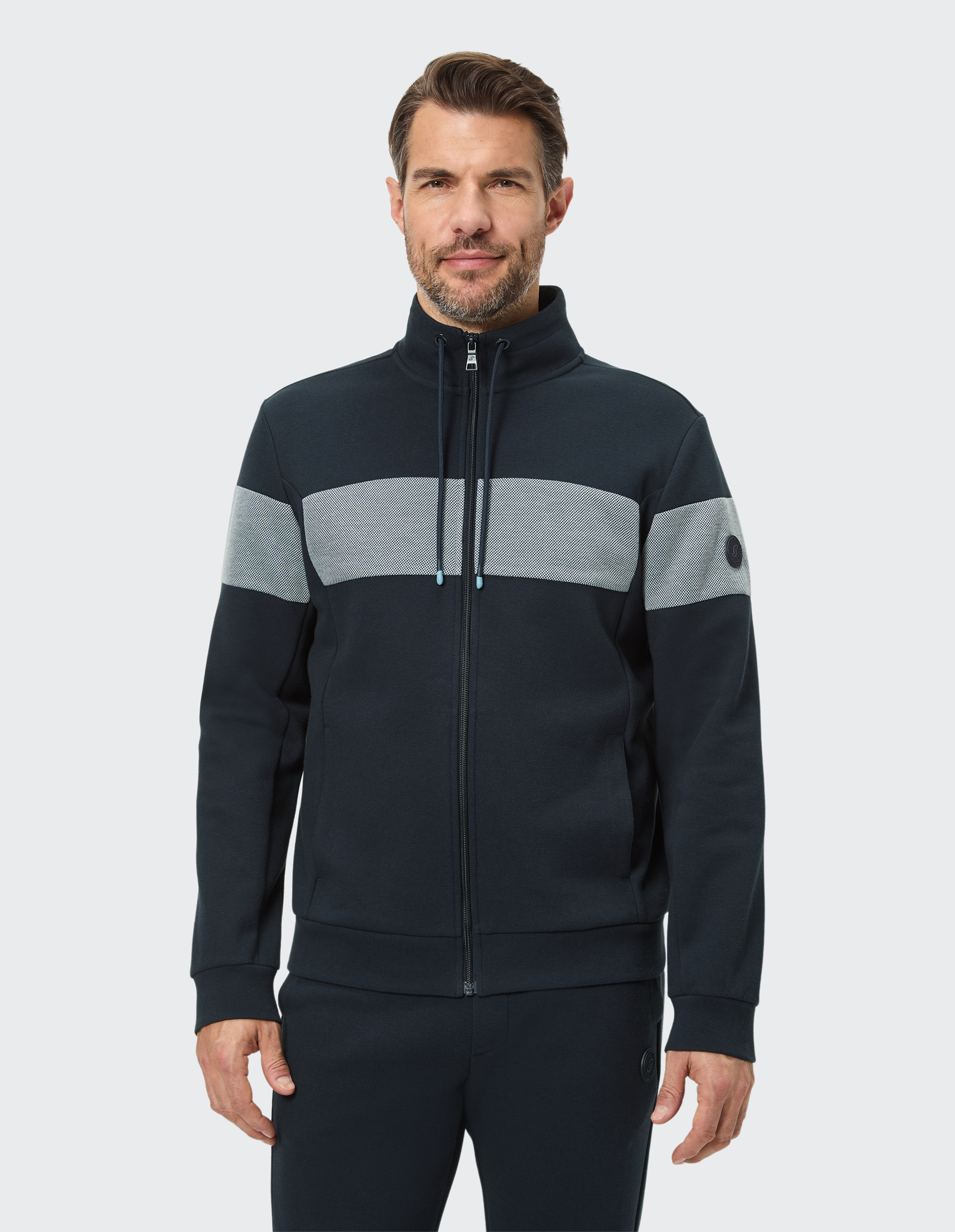 Ein Mann trägt eine dunkelblaue Jacke mit grauem Streifen, Reißverschluss und Kragen, dazu passende dunkelblaue Hose.