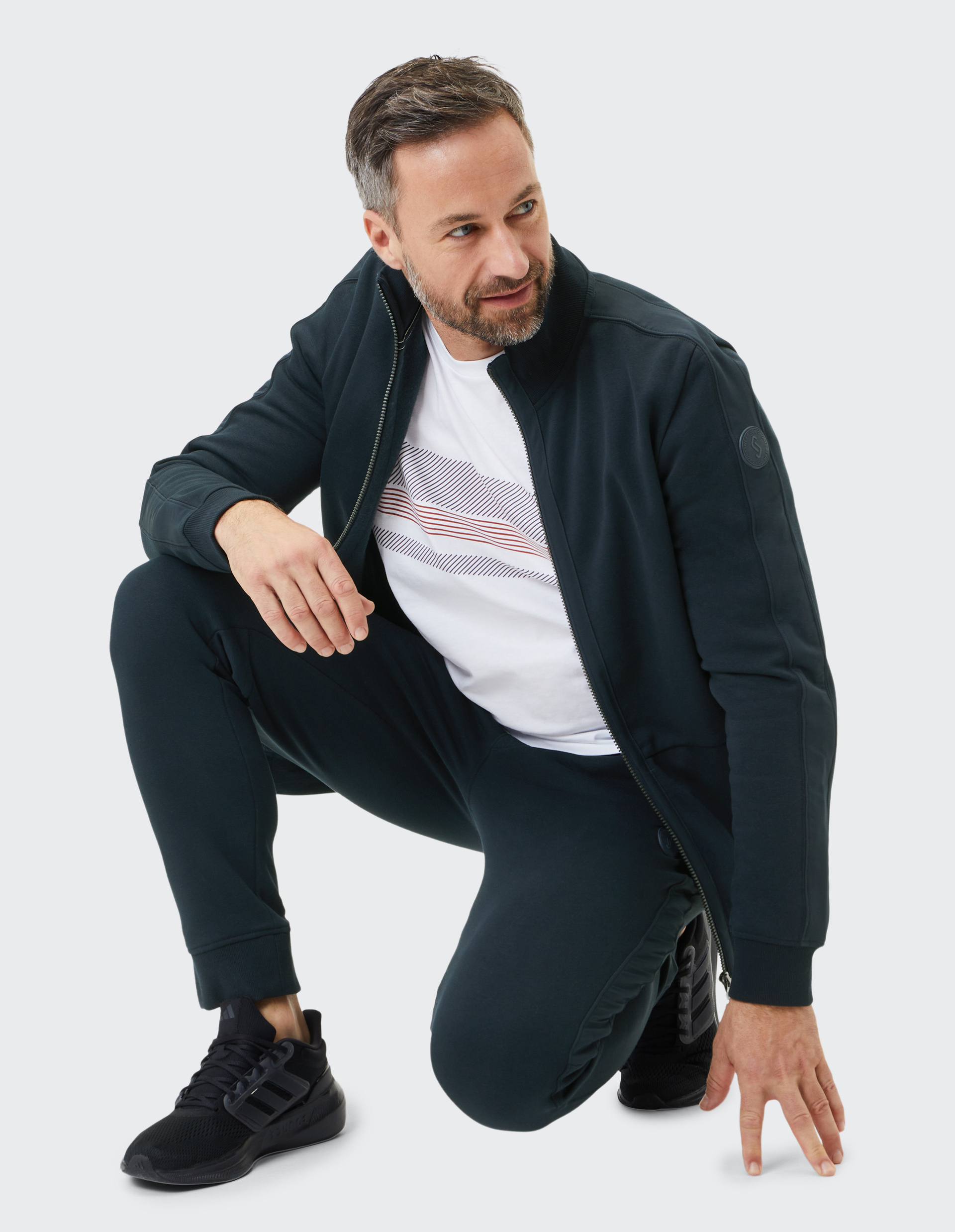 Mann in schwarzem Trainingsanzug und weißen T-Shirt kniet, trägt Sportschuhe, blickt seitlich, vor hellem Hintergrund.