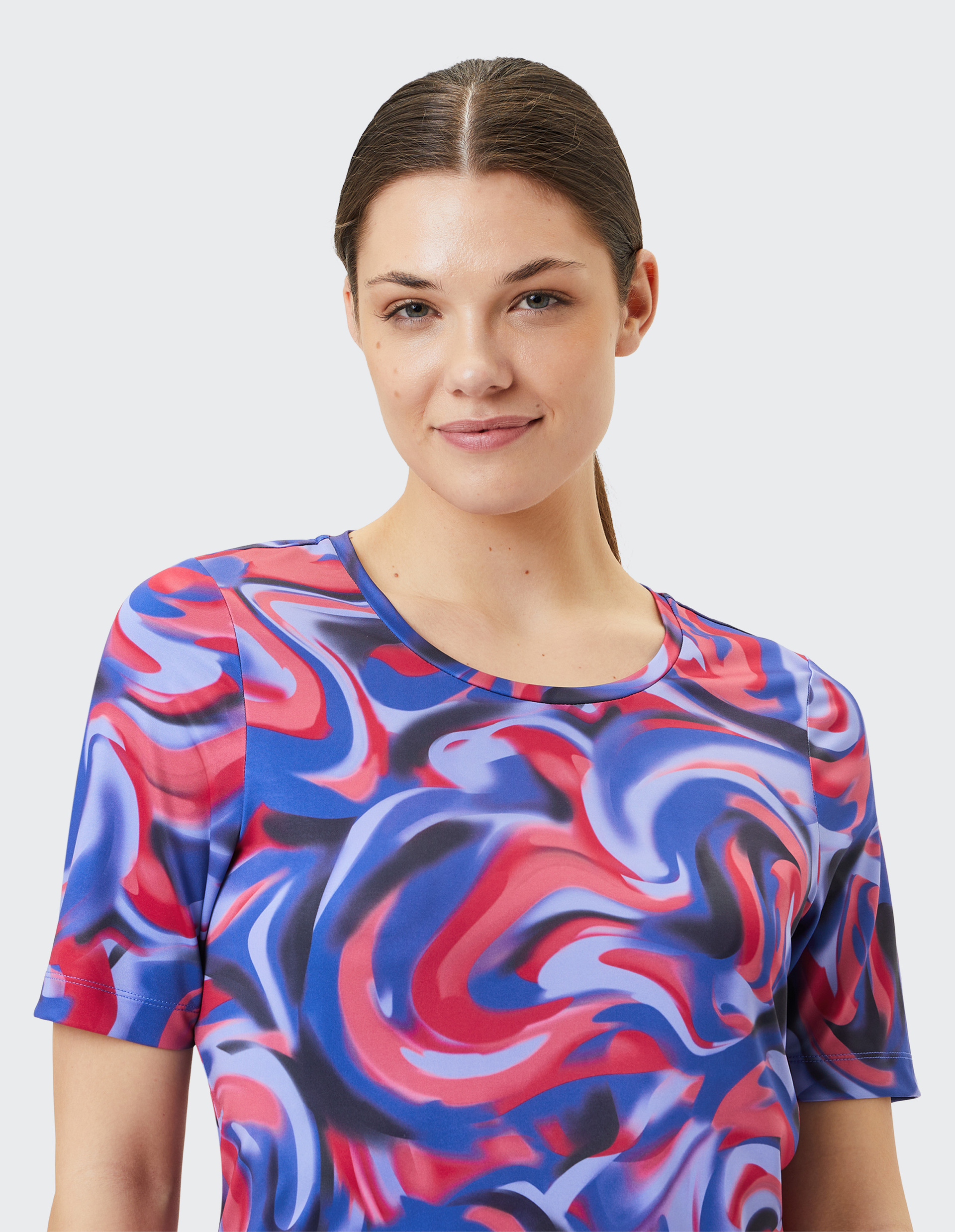 Frau mit braunen Haaren trägt buntes T-Shirt mit dynamischem roten, blauen und lila Wirbelmuster vor hellem Hintergrund.