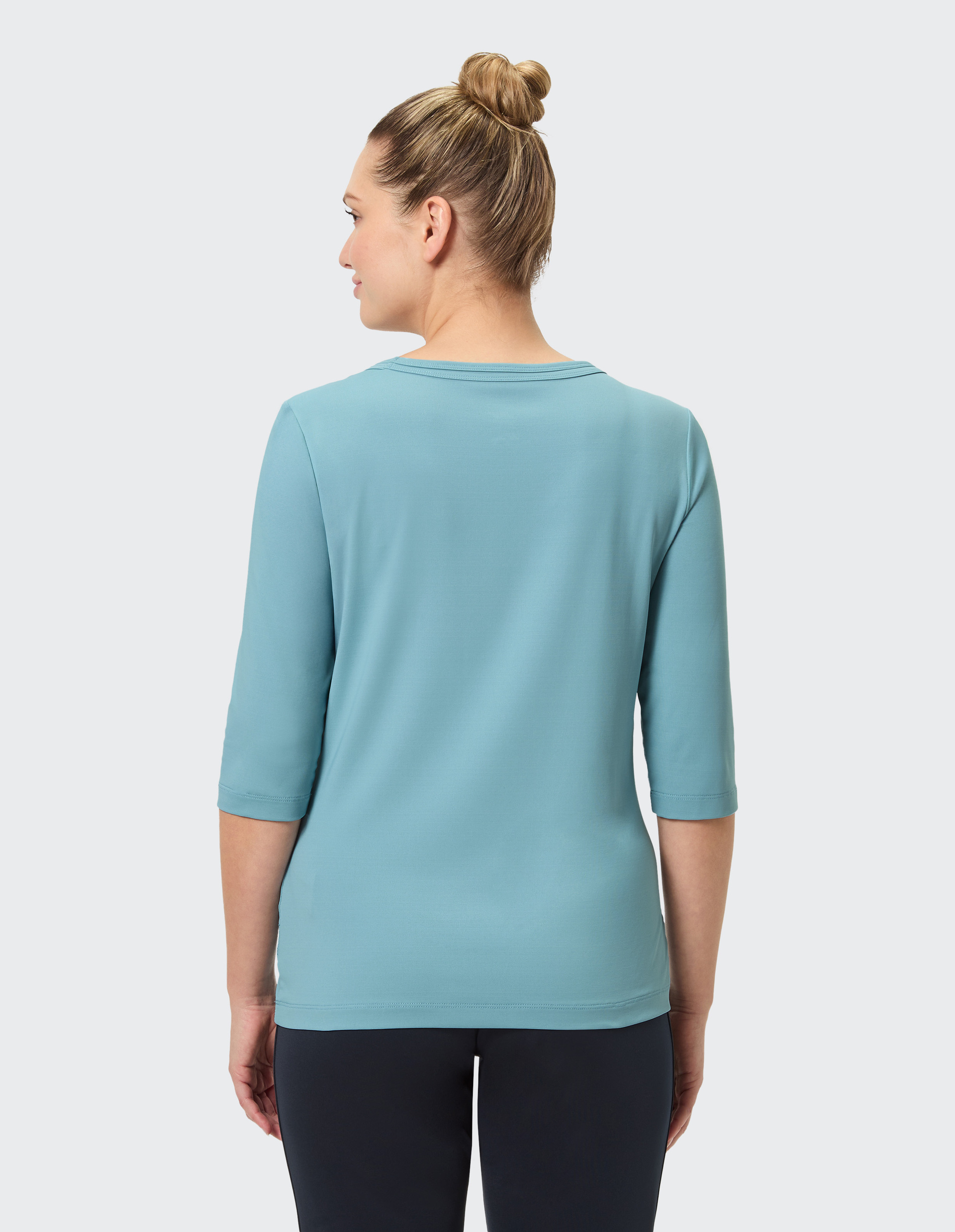Eine Frau in einem hellblauen Oberteil mit Dreiviertelärmeln steht mit dem Rücken zur Kamera, ihre Haare sind zu einem Dutt gebunden.