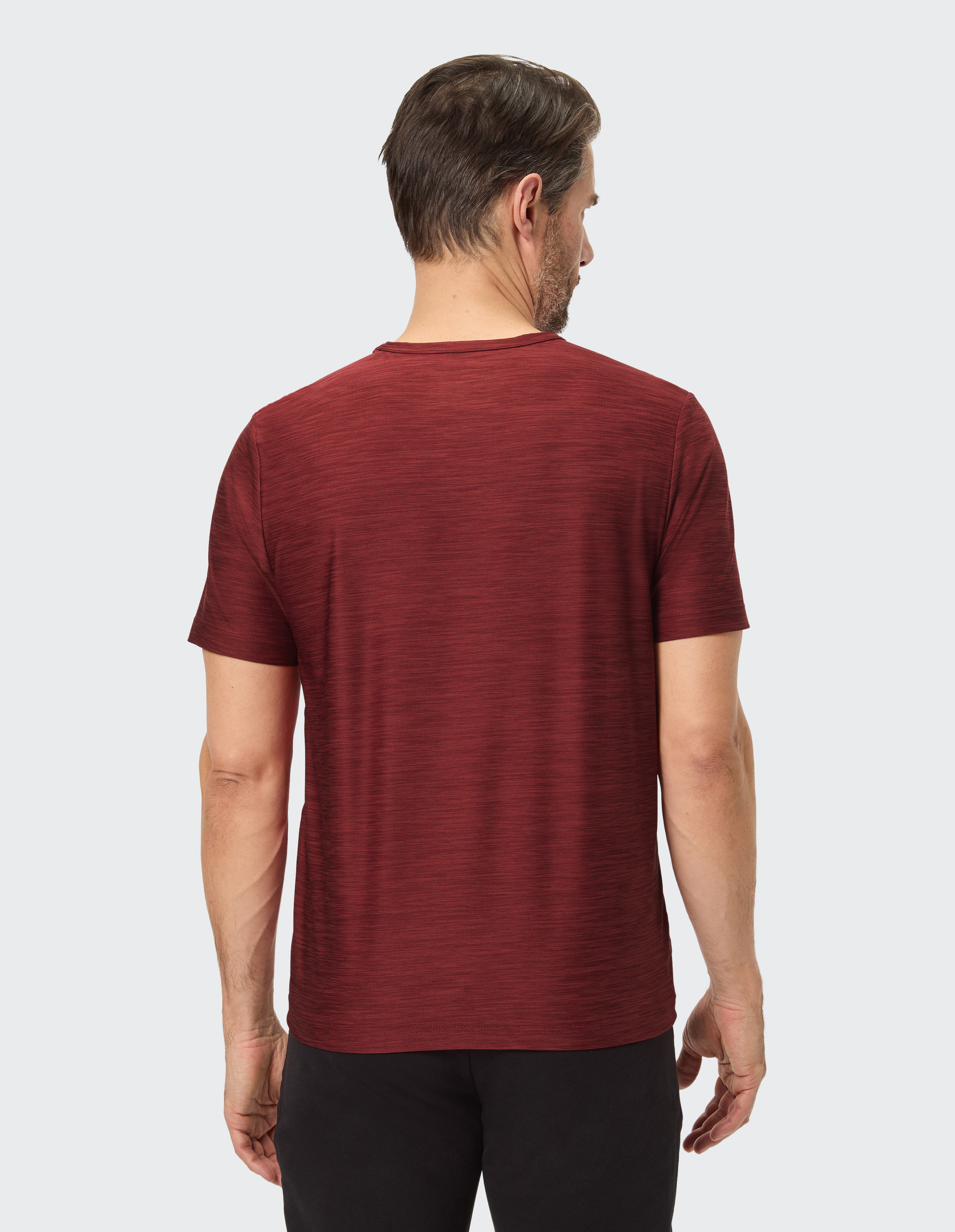 Ein Mann in einem roten T-Shirt steht mit dem Rücken zur Kamera, während er nach rechts schaut; Hintergrund ist einfarbig.