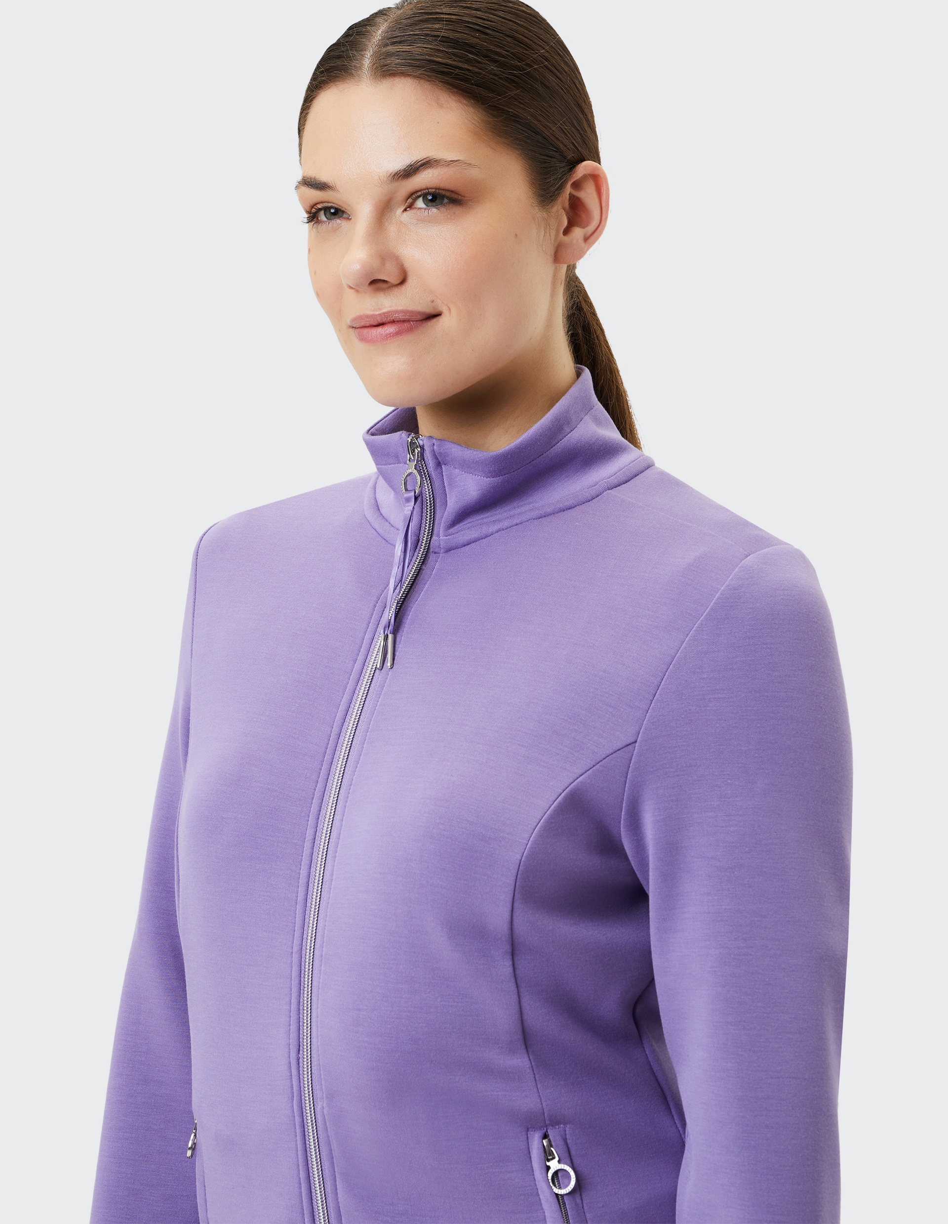 Eine Frau mit zurückgebundenem Haar trägt eine lila Jacke mit Reißverschluss und Stehkragen vor hellem Hintergrund.