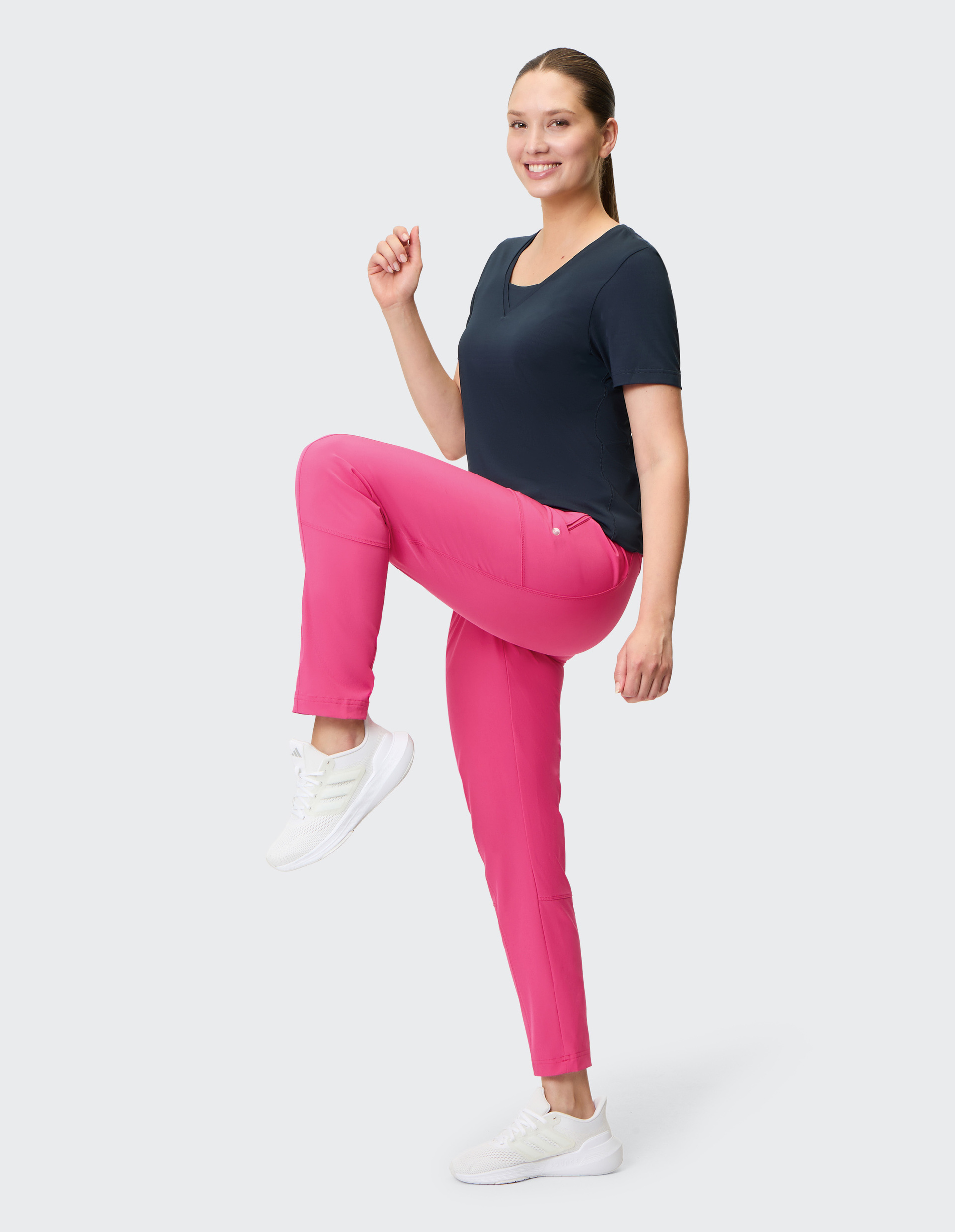 Eine lächelnde Frau in schwarzem T-Shirt und pinker Hose hebt ein Bein, als würde sie trainieren, und trägt weiße Sneakers.